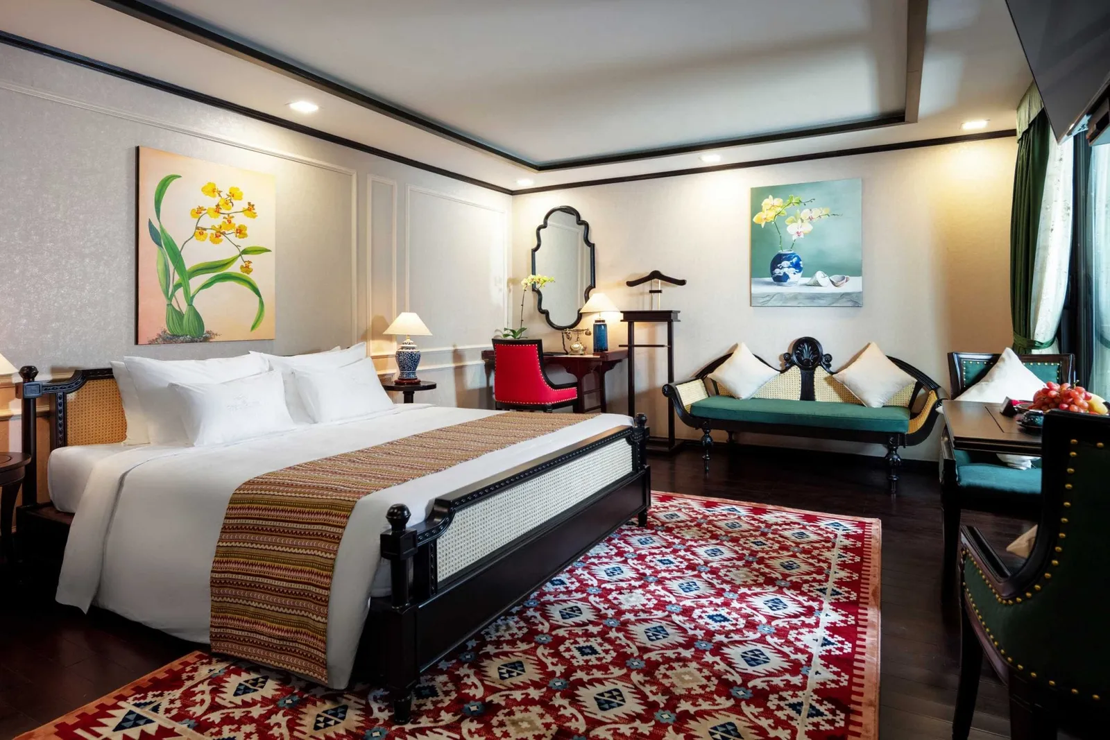A luxurious cabin interior with a large bed, decorative rug, seating area, ornate mirror, floral artwork, and dark wood furniture.