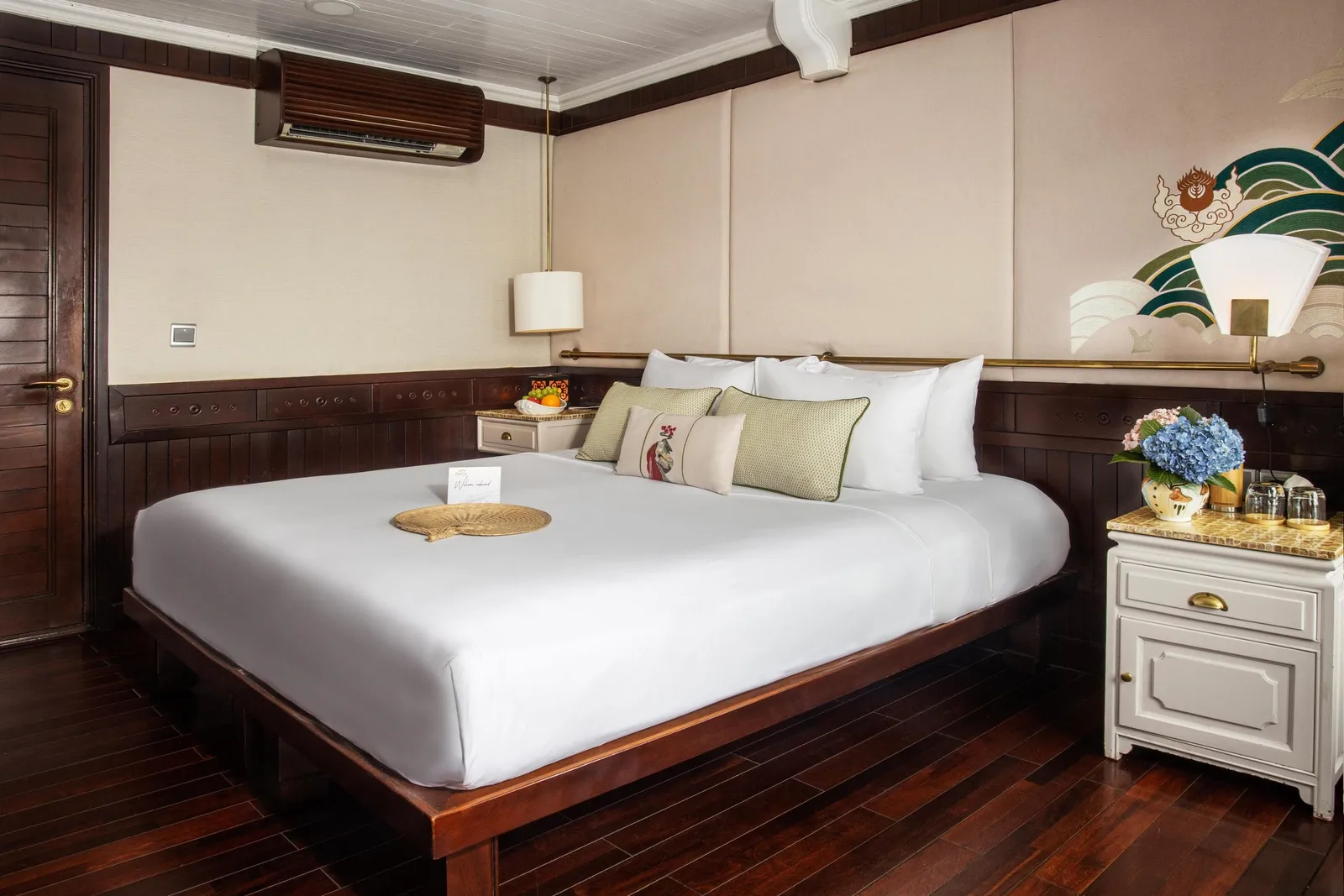 A cruise ship cabin interior with a large bed, dark wood paneling, white bedding, decorative pillows, and a white nightstand with flowers.
