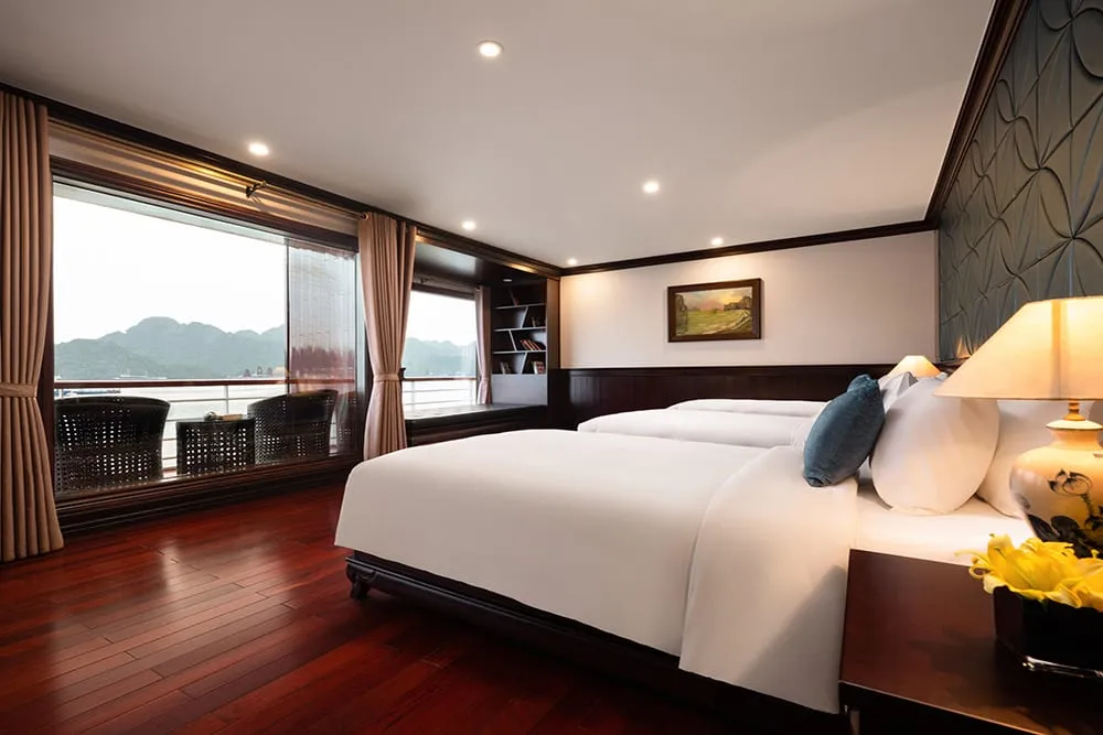 A cabin interior with two beds, dark wood flooring, and large windows showing limestone karst islands across the water.