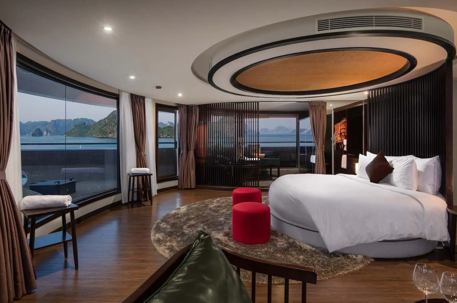 A spacious cruise cabin with a round white bed, curved floor-to-ceiling windows, an oval ceiling feature and red stools, with karst islands visible over the water outside.