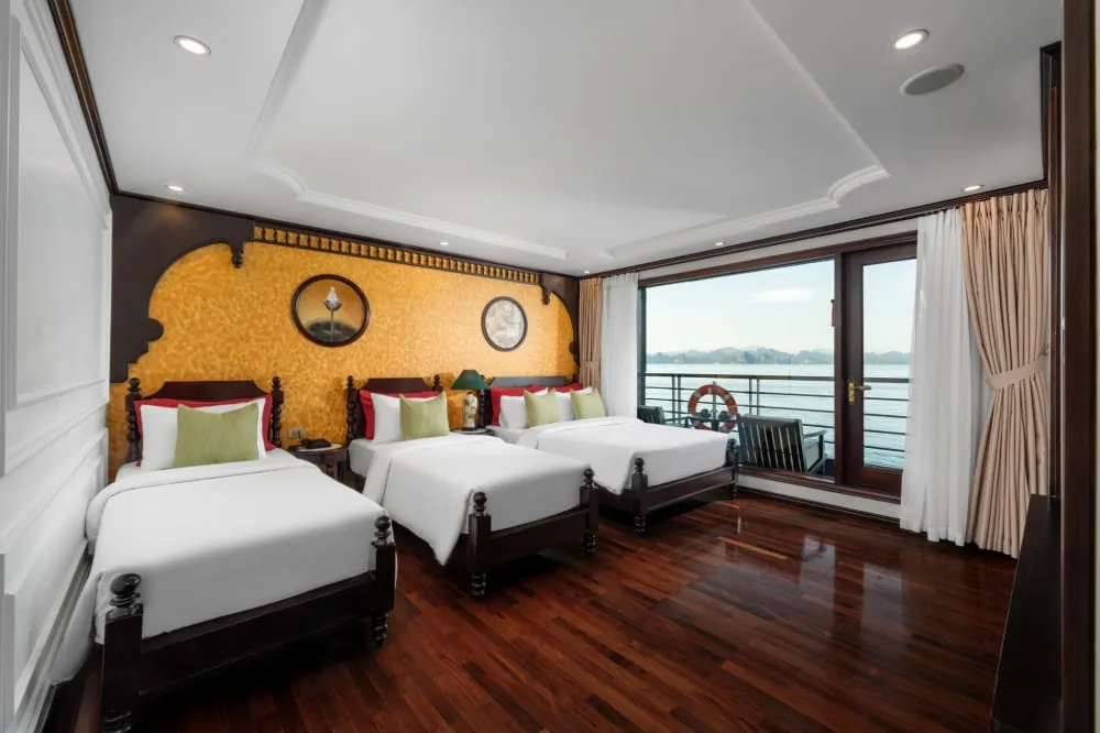 A cruise boat cabin interior with three beds, wooden floors, decorative gold textured wall panels, and large windows showing karst formations and water outside.