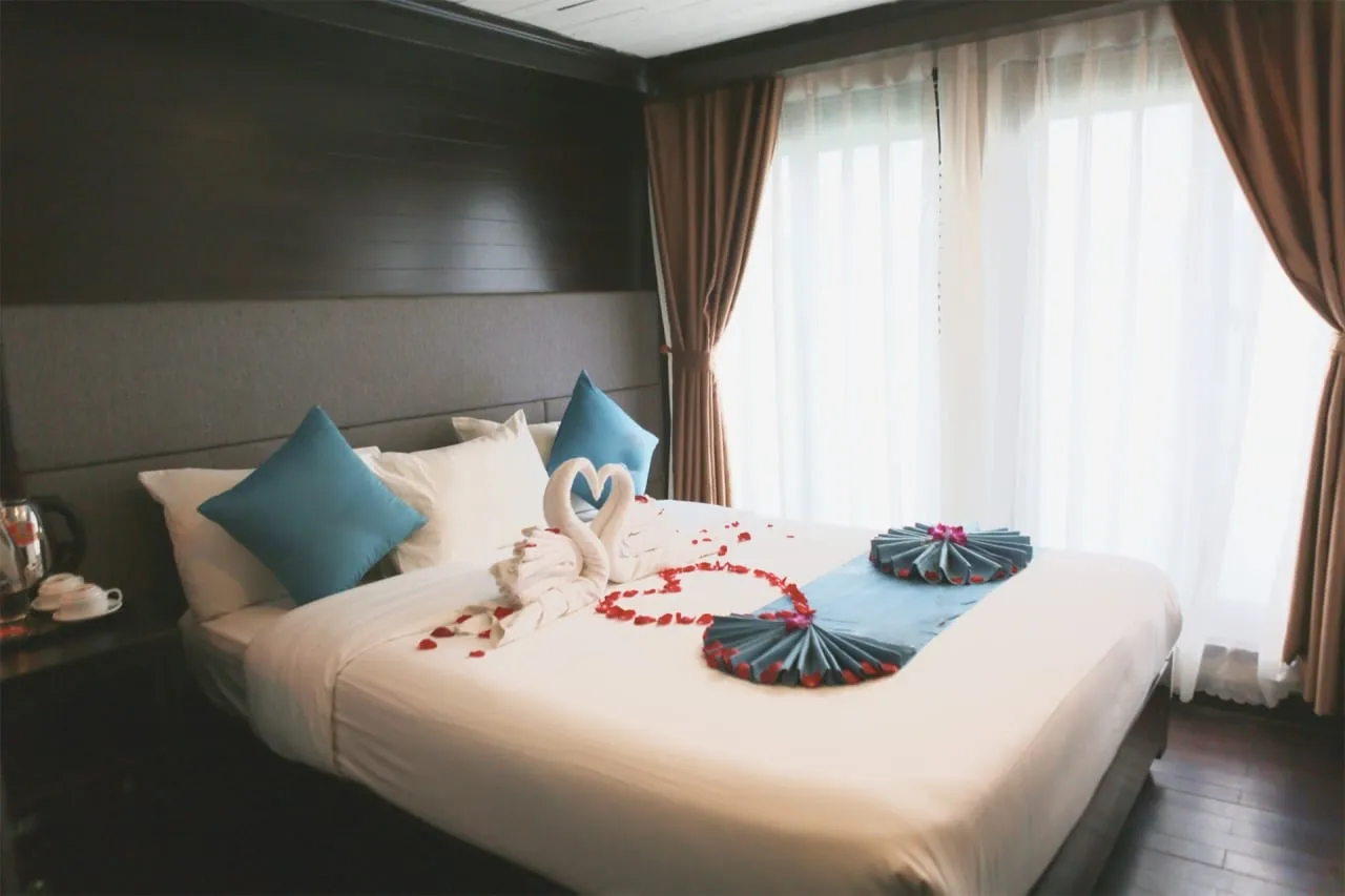 A cabin with a double bed dressed in white linens, towel swans, rose petals in a heart shape, teal cushions and folded fans, with sheer and brown curtains over a bright window.