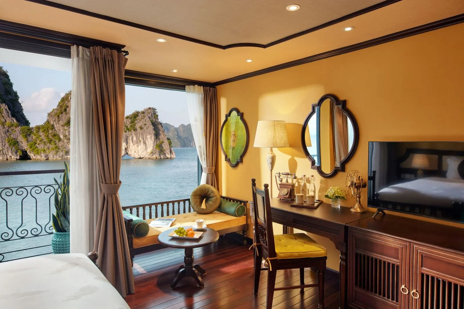 A yellow-walled wood-trimmed cabin with a window-side daybed, desk with vintage phone, mirrors and TV, opening onto a balcony overlooking calm water and limestone karst islands.