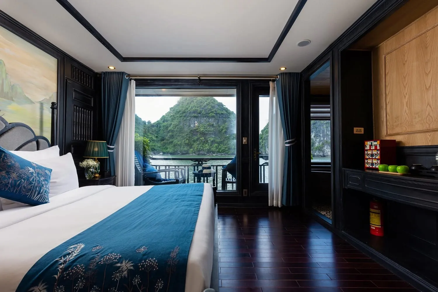 Interior view of a cruise cabin with a bed, dark wood finishes, and a balcony door framing a limestone karst formation in the bay beyond.