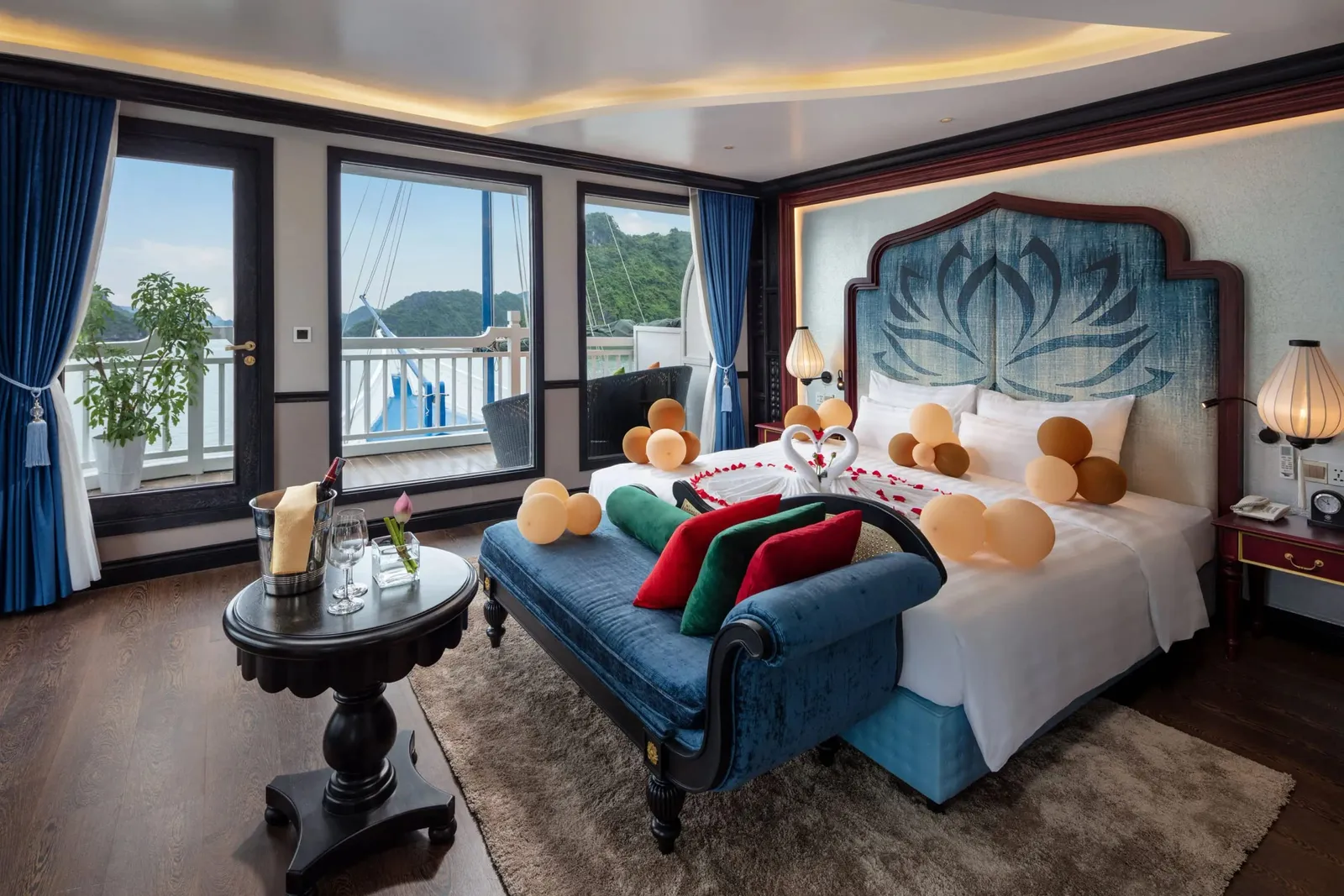 A cabin with a decorated bed (balloons, towel swans, rose petals), a blue chaise, an ice bucket on a side table, and windows and a door opening onto a balcony with limestone karst hills beyond.