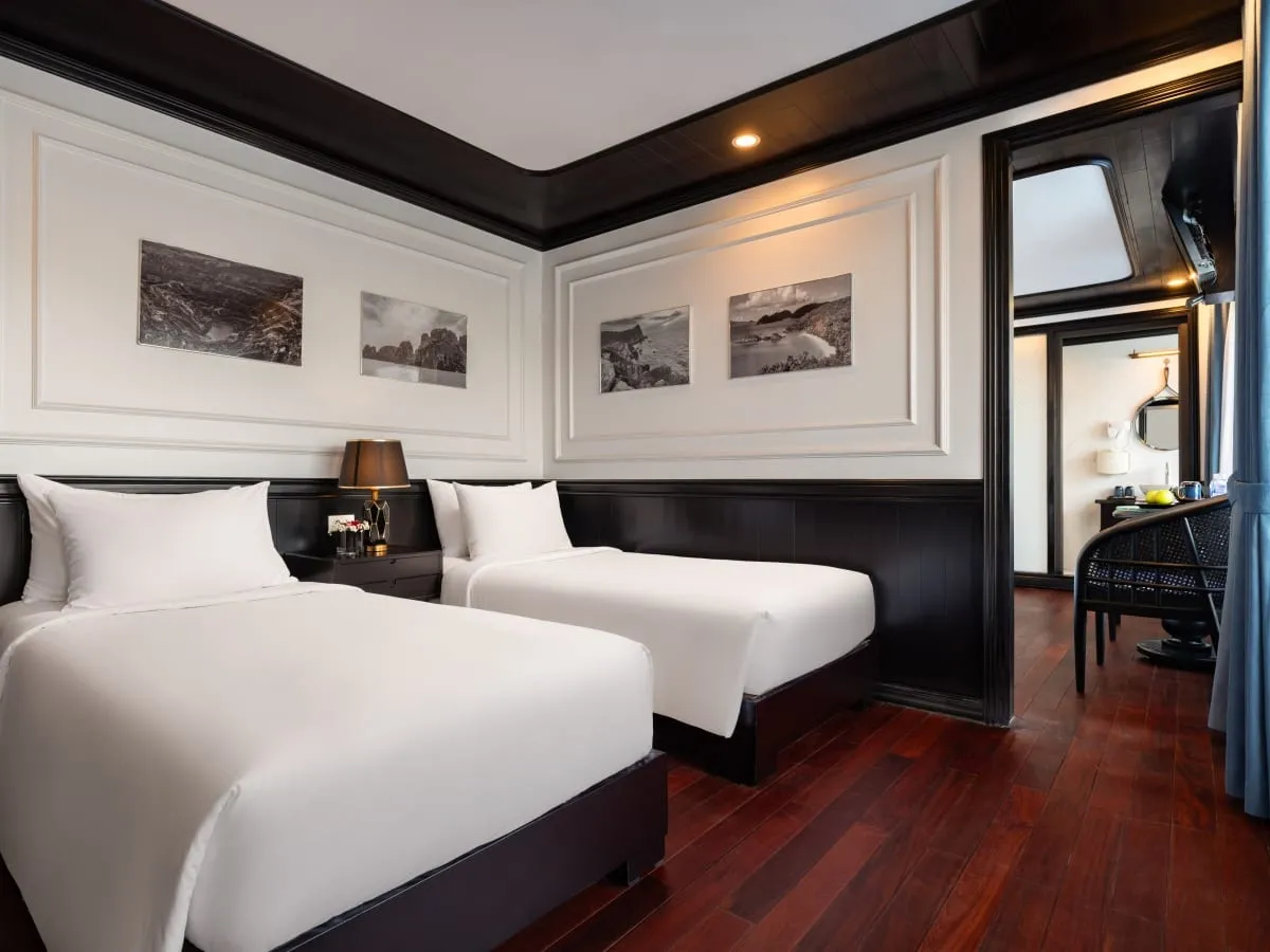 A twin-bed cabin with white bedding, dark wood paneling and floor, framed black-and-white coastal photos on the wall, a bedside lamp, and a doorway leading to a small desk area and a porthole-style window.