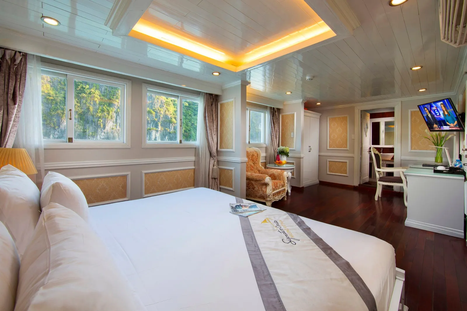 A cabin with a large white bed, a bed runner bearing a logo, a dark wood floor, an armchair, a desk with a wall-mounted TV, and windows looking out on forested limestone karst cliffs.