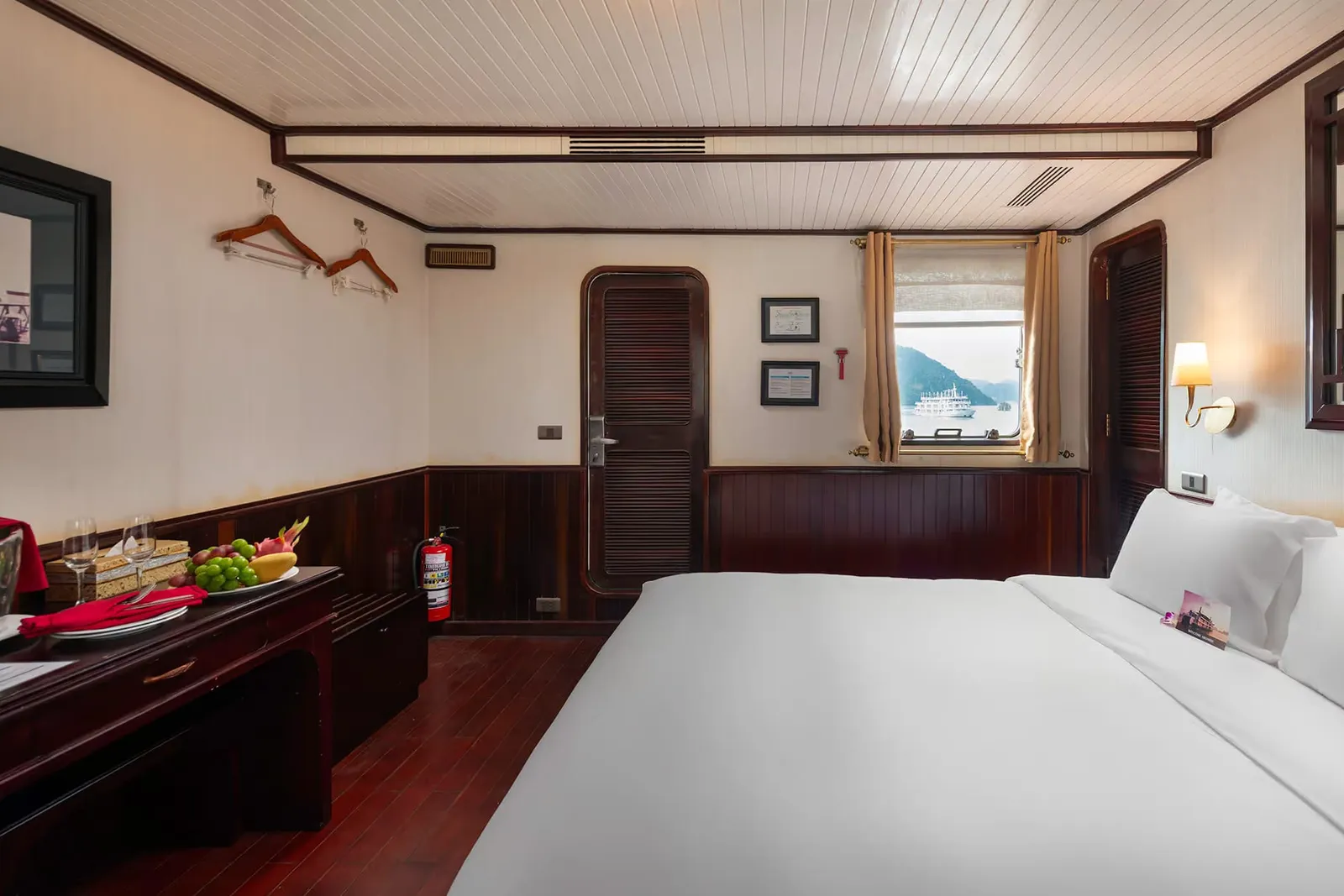 A wood-paneled cruise cabin with a large white bed, a desk holding a fruit plate and wine glasses, a fire extinguisher, and a window showing a white boat and karst hills on the water.