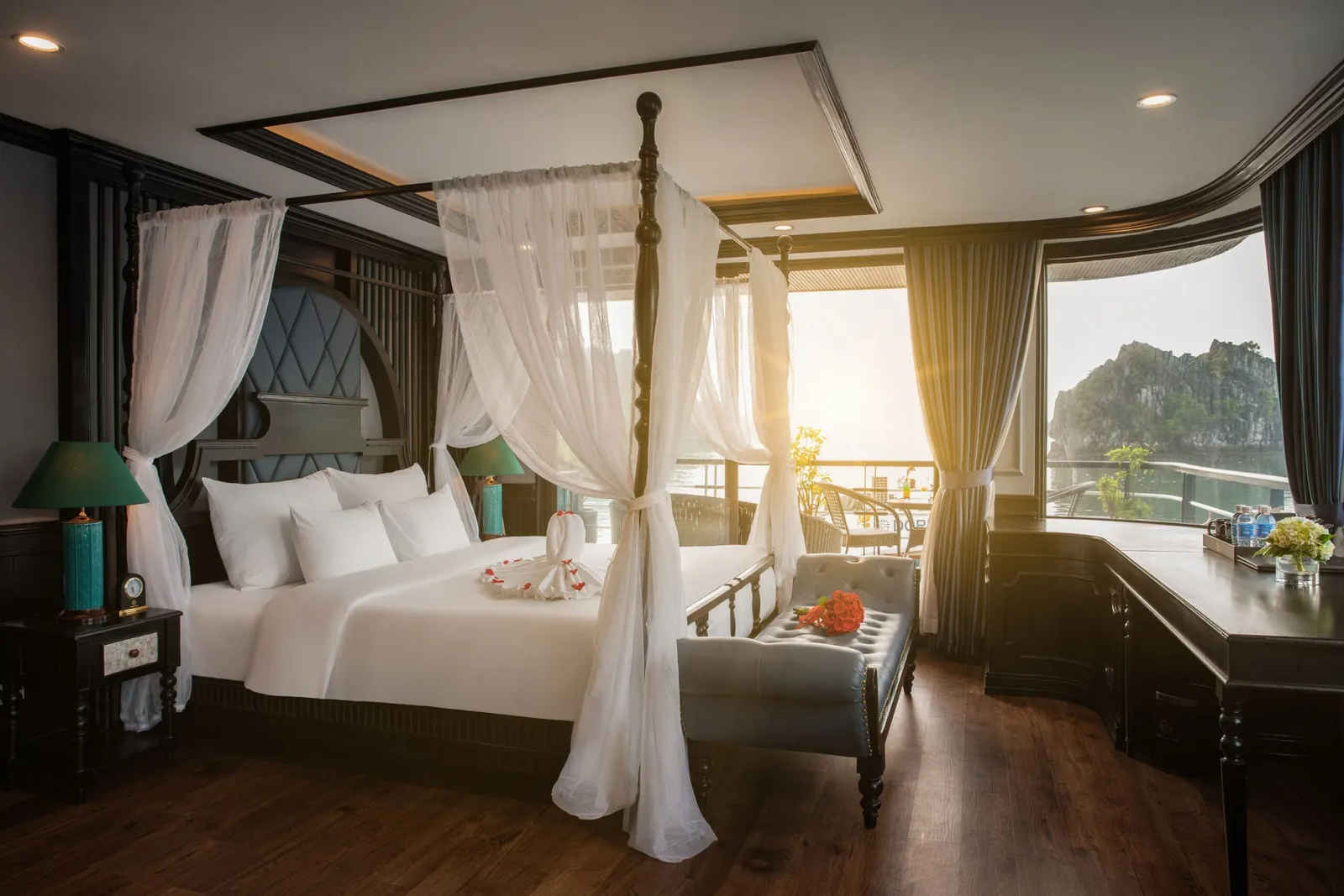 A luxury cabin interior with a canopy bed and large windows showing karst limestone formations and water typical of Ha Long Bay.