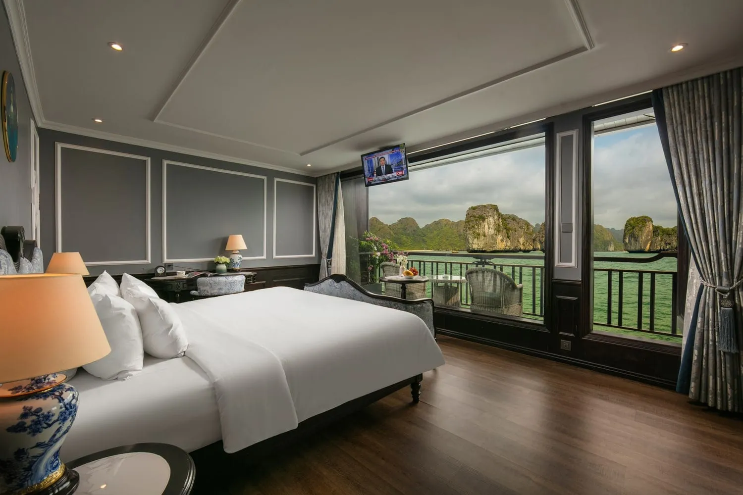 A luxury cabin interior with a white bed, wooden floor, and large windows showing limestone karsts and green water outside.