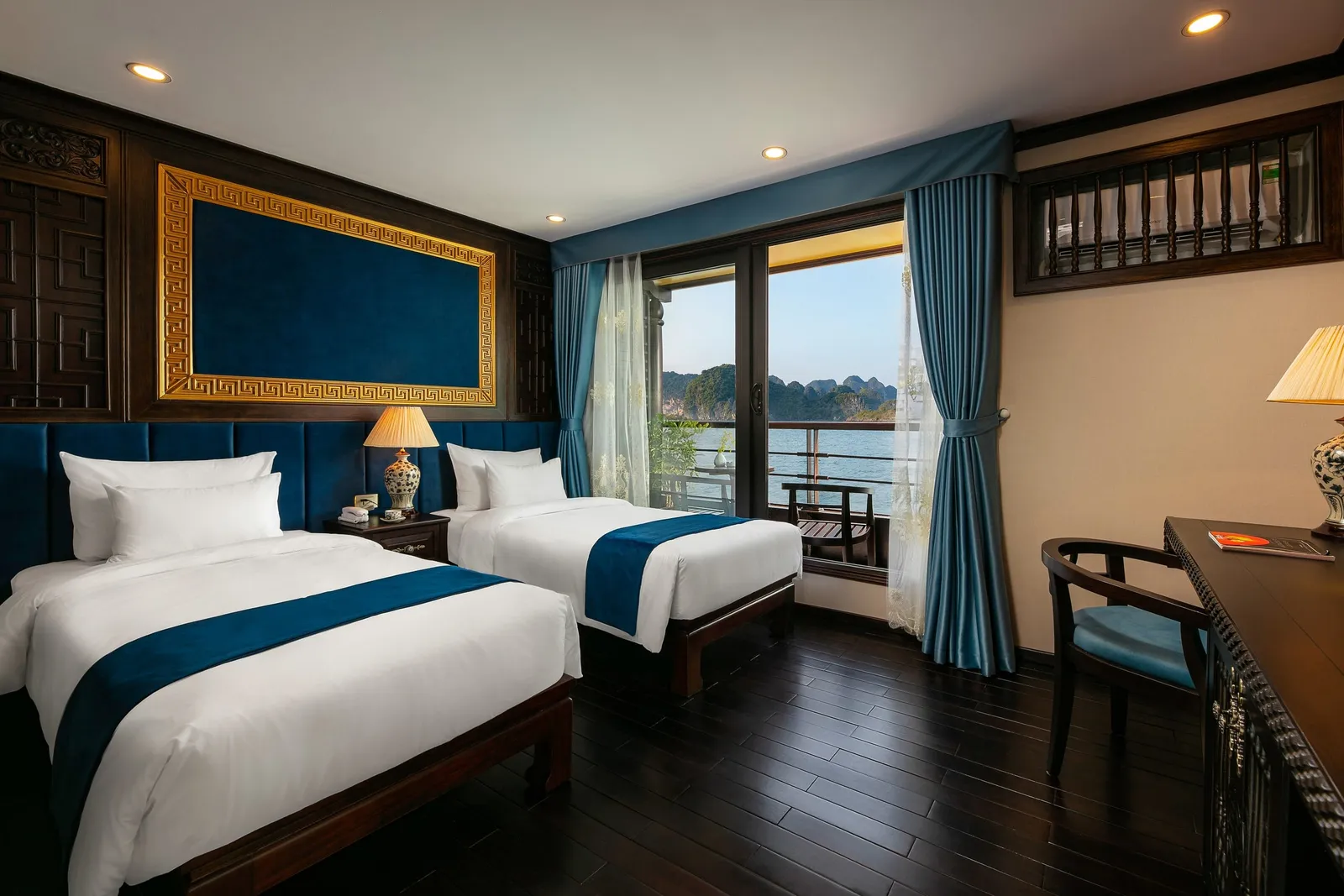 A twin-bed cabin with dark wood floor, blue and gold decor and a balcony door looking out onto water and limestone karst islands.