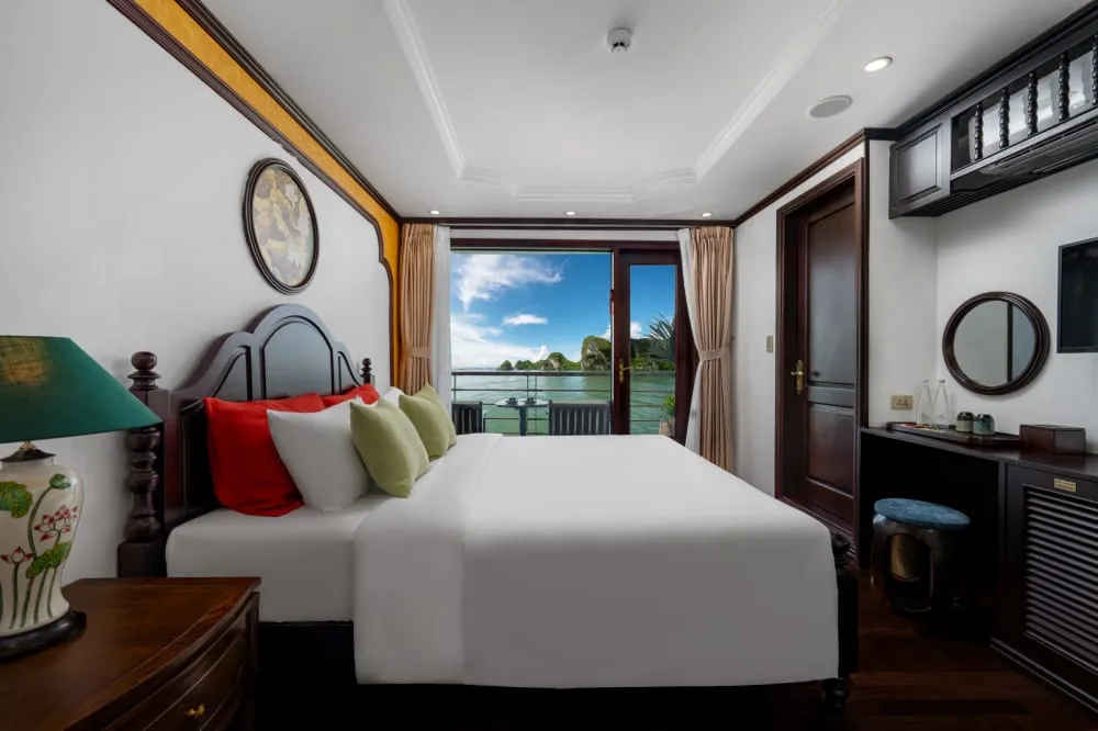 A boat cabin interior with a bed, dark wood furniture, and large windows showing limestone karsts and water outside.