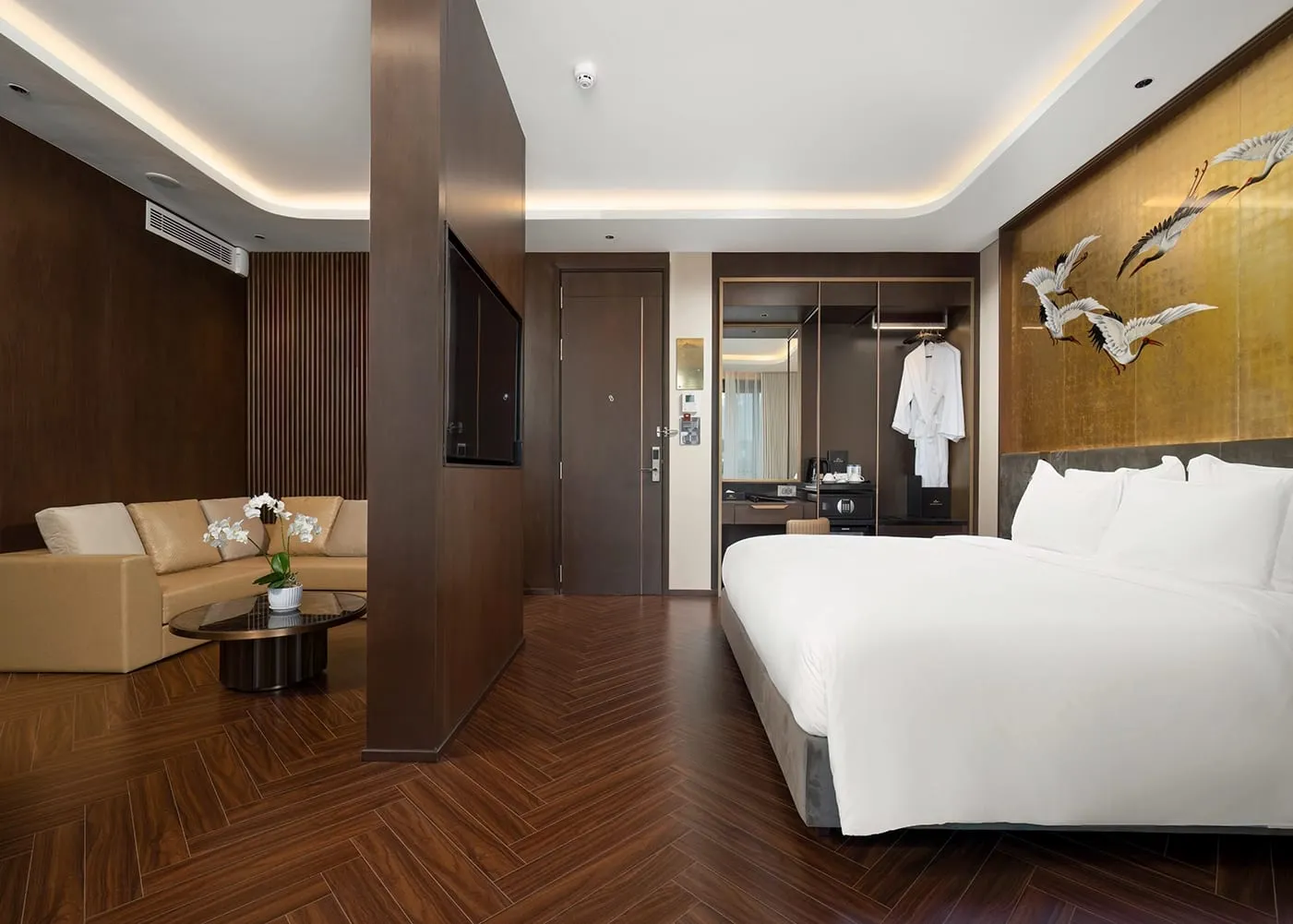 A luxury cruise cabin with a double bed, herringbone wood-effect floor, leather sofa seating area, wardrobe with hanging bathrobe, and a decorative gold-panel mural of cranes on the wall.