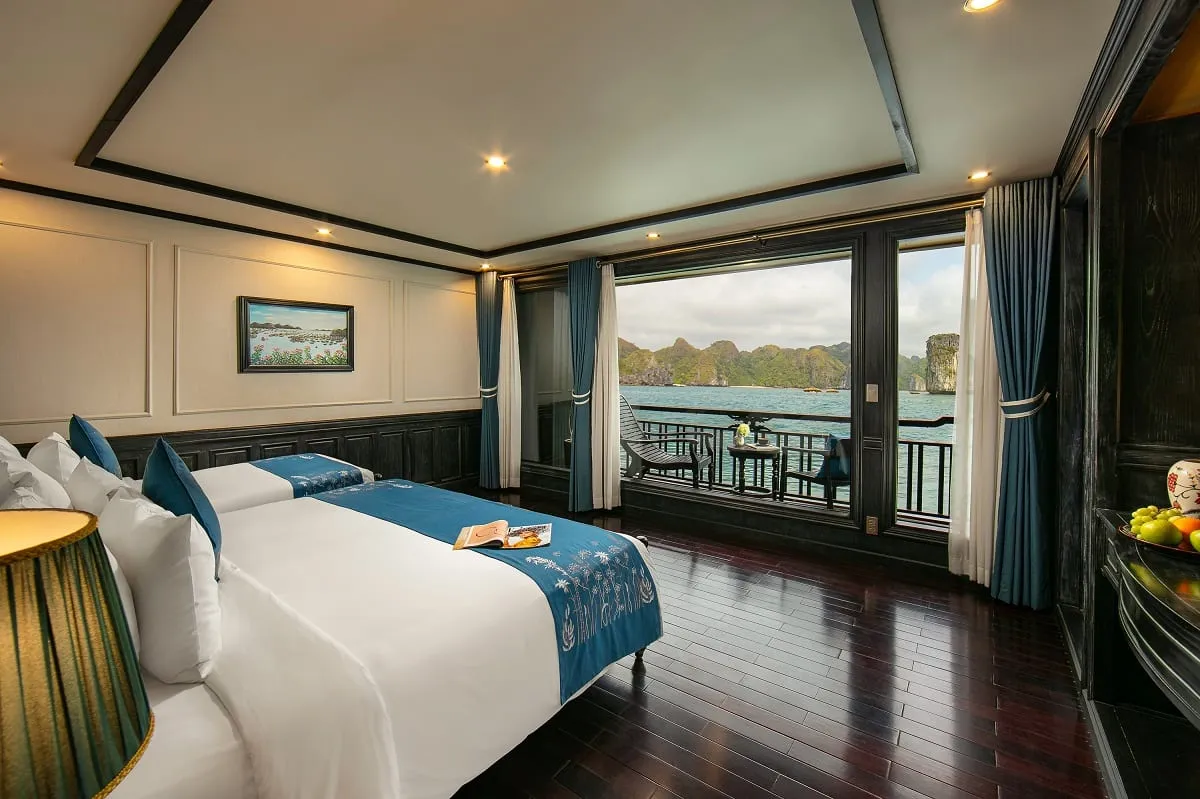A cruise cabin with two white beds, dark wood floors, and large windows opening to a balcony with limestone karsts and turquoise water visible outside.