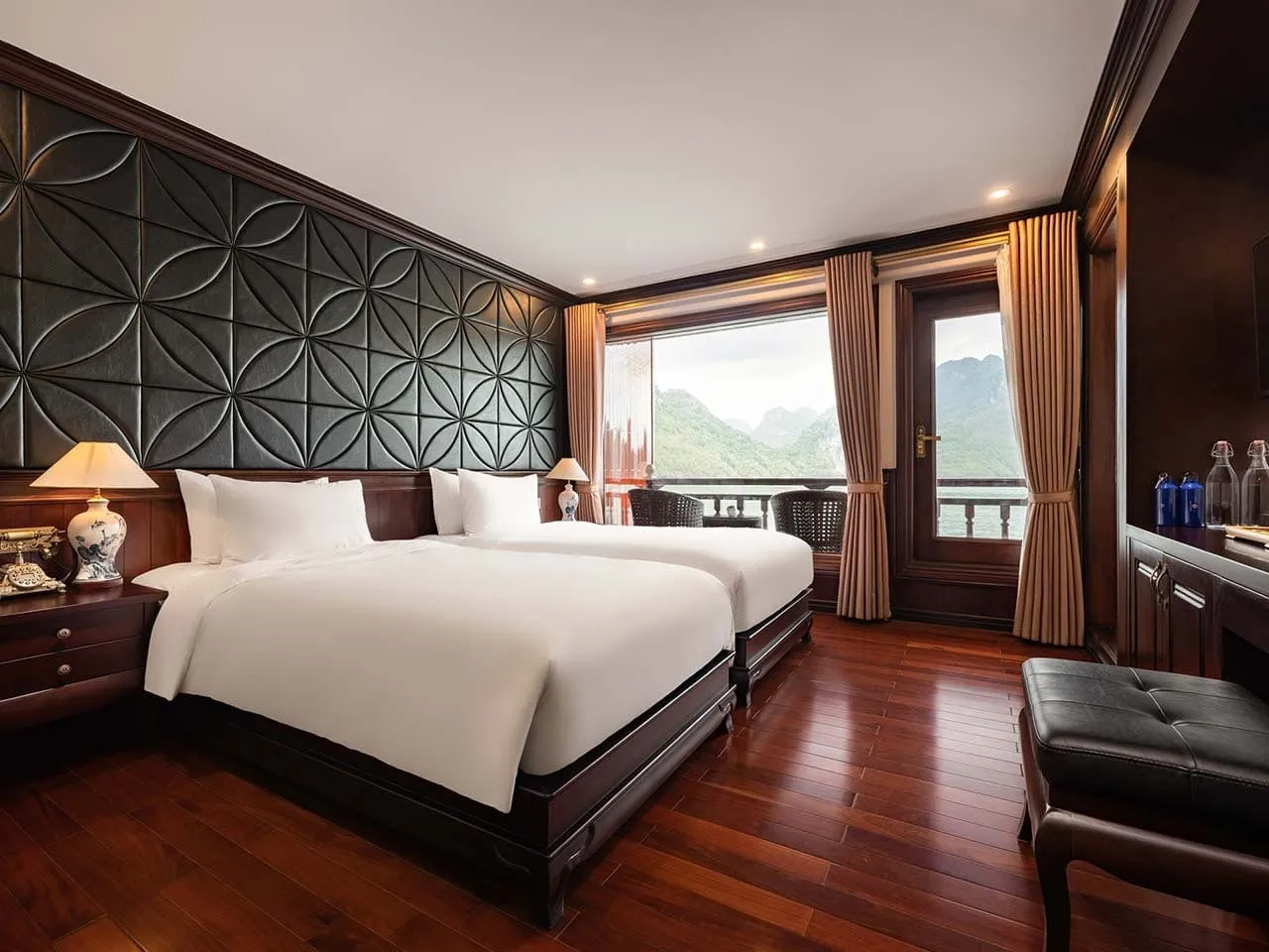 A cabin with two twin beds in white linen, dark wood floor, a quilted dark wall panel, and a glass door opening to a balcony with a view of green karst limestone hills.