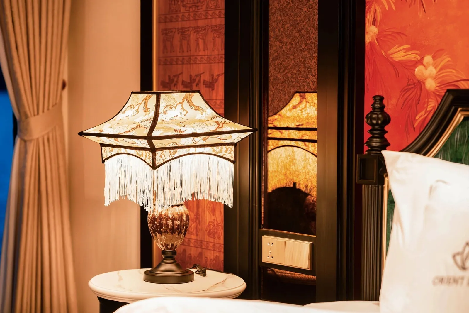 A lit pagoda-style fringed table lamp on a marble bedside table in a warmly decorated cabin, with a curtain, ornate dark wood headboard, and a white pillow embroidered with a logo and partial text reading 'ORIENT'.