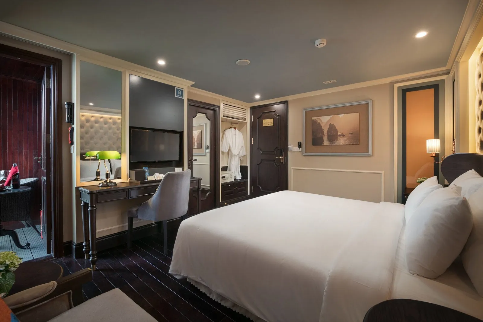 A cabin with a white-bedded double bed, dark wood floor and doors, a desk with TV and green banker's lamp, a wardrobe with a bathrobe, a framed Ha Long Bay karst painting, and a door opening to a balcony with a small table.