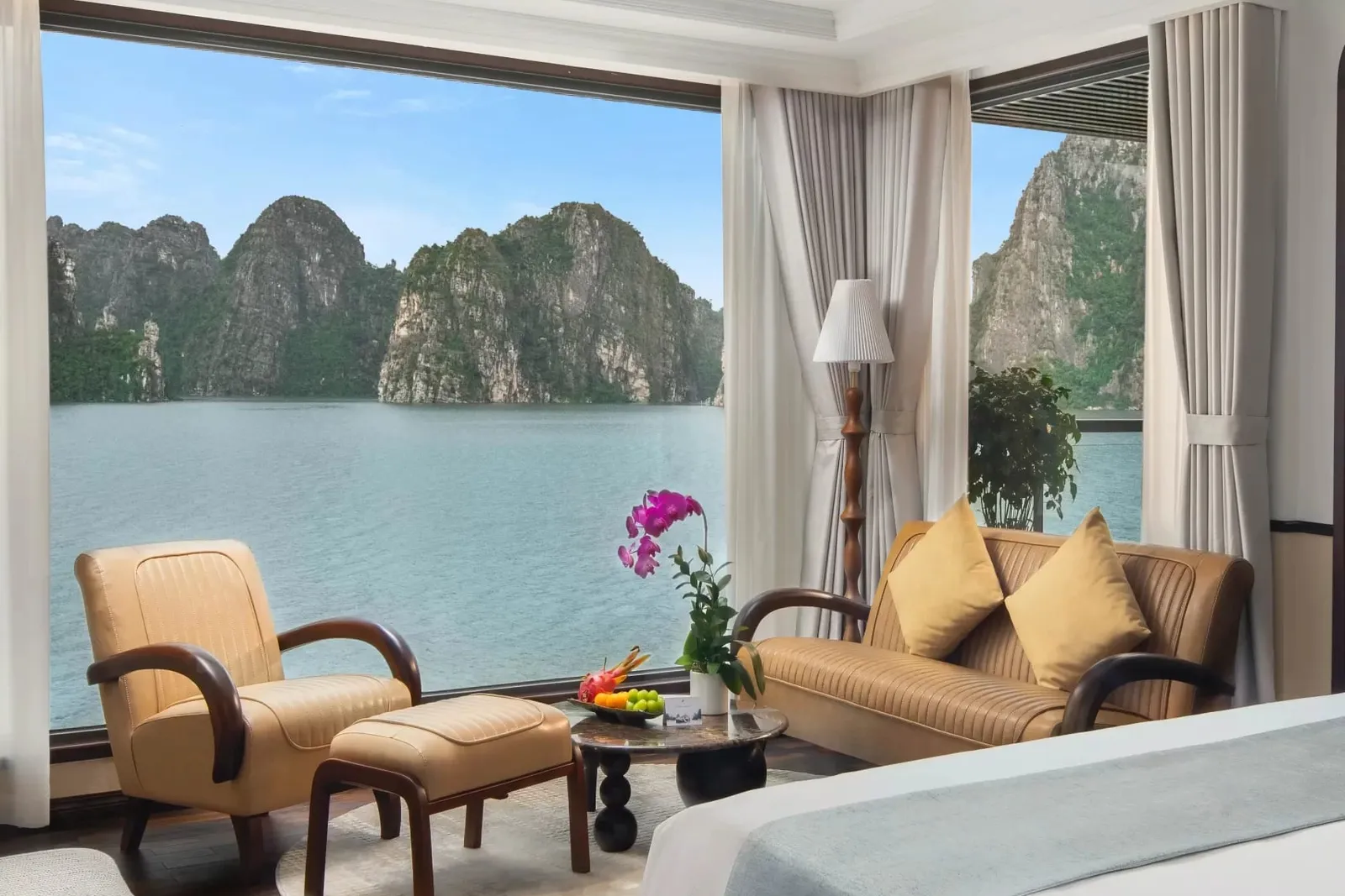 A luxury cabin interior with a bed, seating area, and large windows showing limestone karsts and calm water.