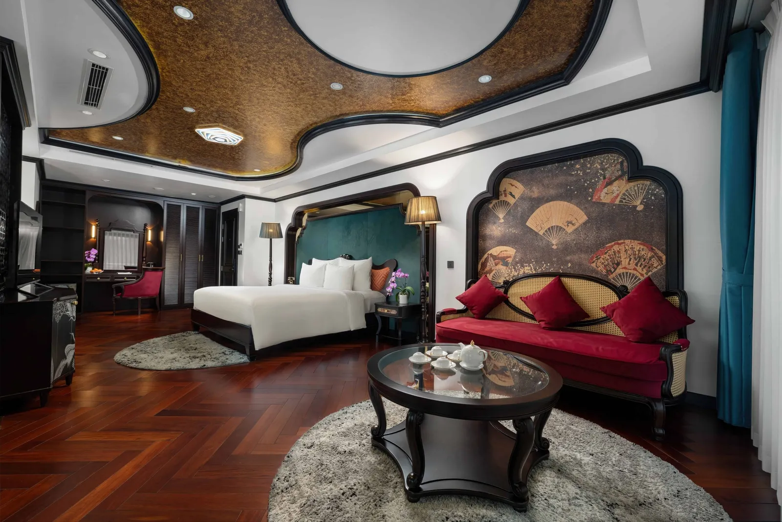 A luxurious cabin interior with a white double bed, red velvet sofa under a fan-patterned panel, round glass coffee table with tea set, herringbone wood floor and a gold-toned curved ceiling.