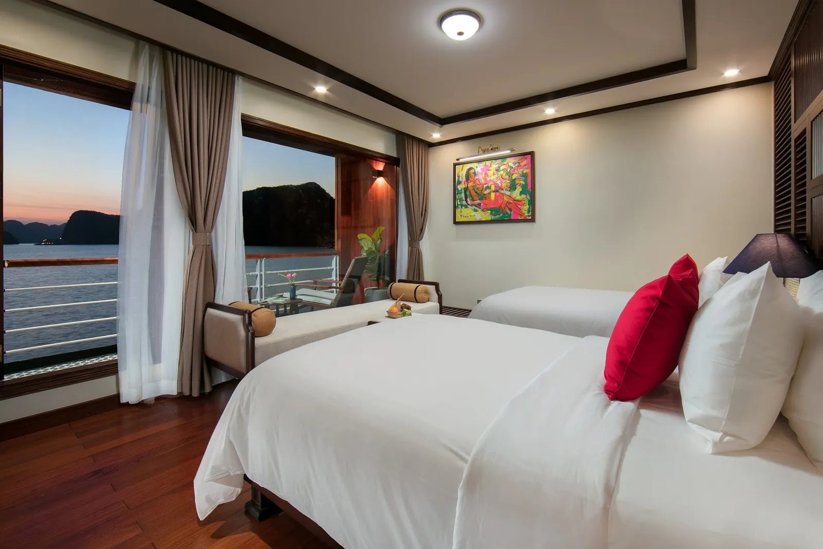 A cruise cabin interior with twin beds, wooden floors, and large windows showing karst limestone formations in a bay at sunset.