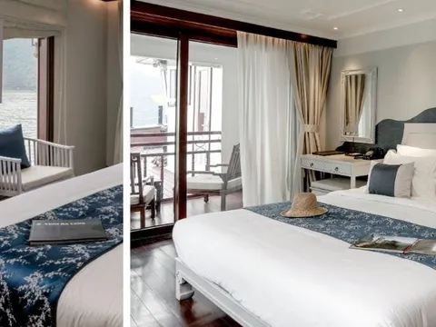 Two side-by-side views of an elegantly furnished cruise cabin with a double bed dressed in blue and white linen, a wall-mounted TV, balcony doors opening onto a water view with karst limestone scenery, and a booklet on the bed reading 'VINH HA LONG'.