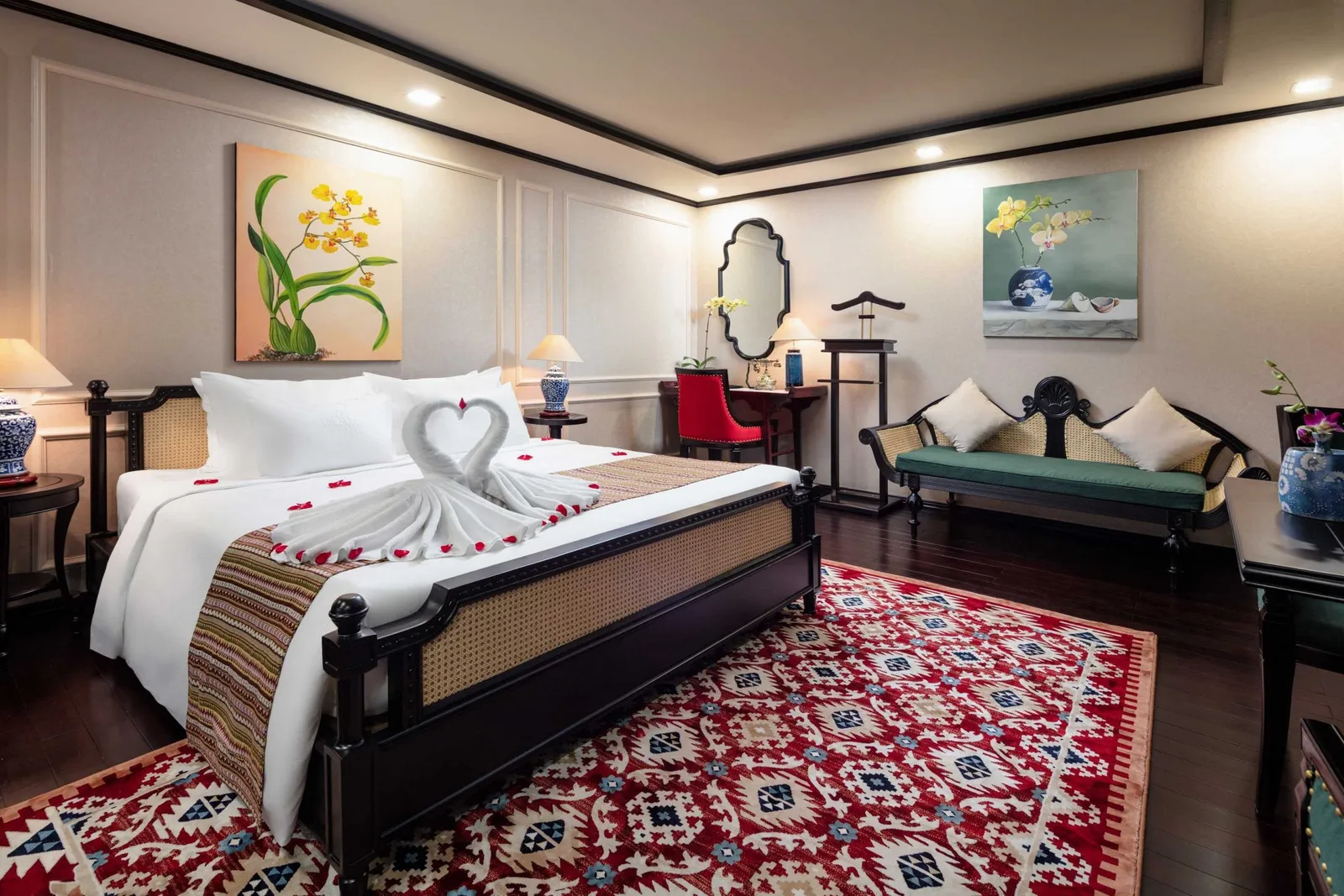 A luxurious cabin interior with a bed decorated with rose petals, dark wood furniture, orchid paintings, and a patterned red carpet.
