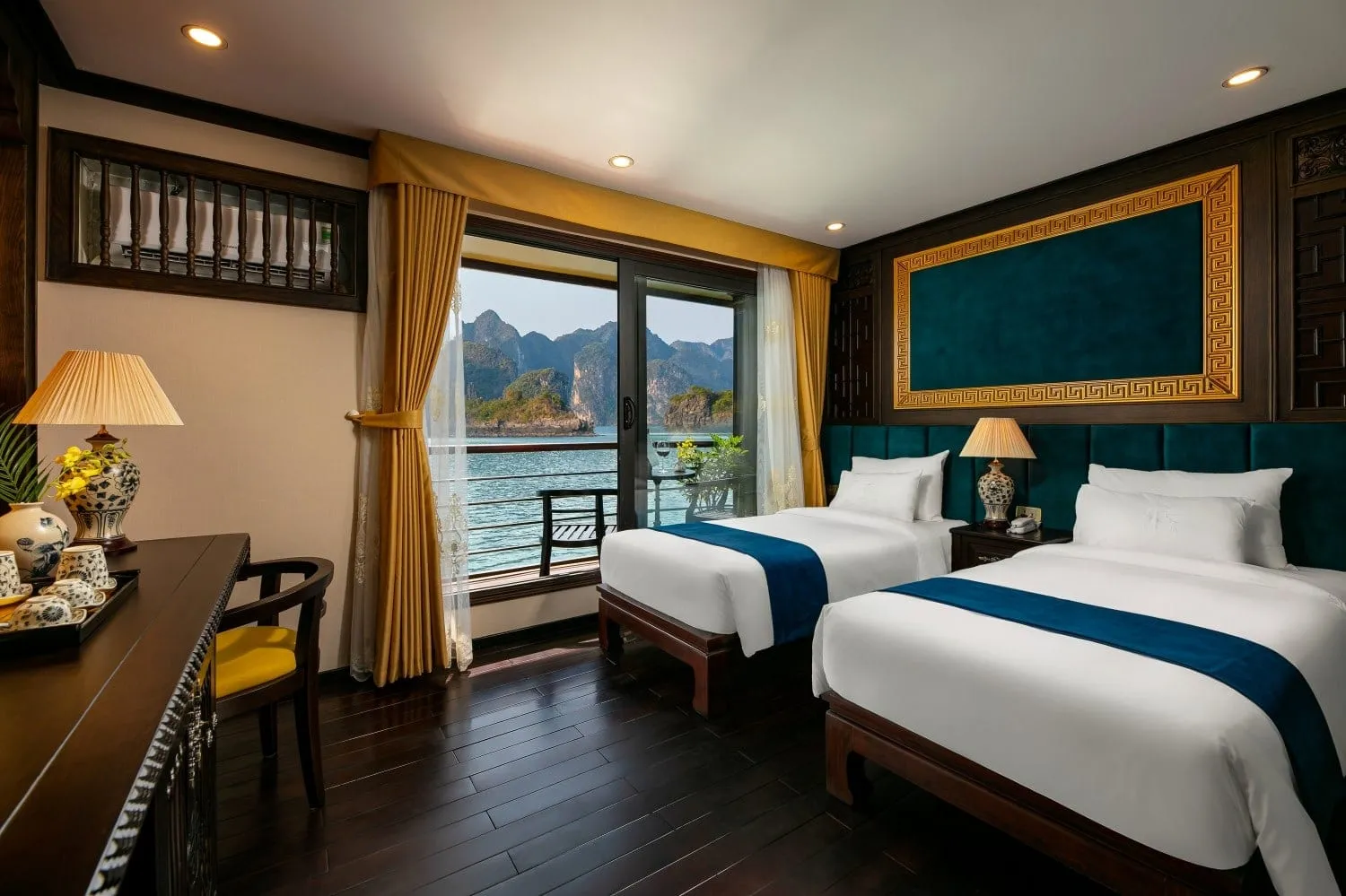 A twin-bed cabin with dark wood floor, teal headboard panels and gold curtains, with a glass door opening to a balcony overlooking water and limestone karst islands.