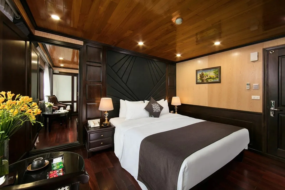 A cruise boat cabin interior with a bed, wooden ceiling, dark wood furniture, yellow flowers, and a view into an adjacent seating area.