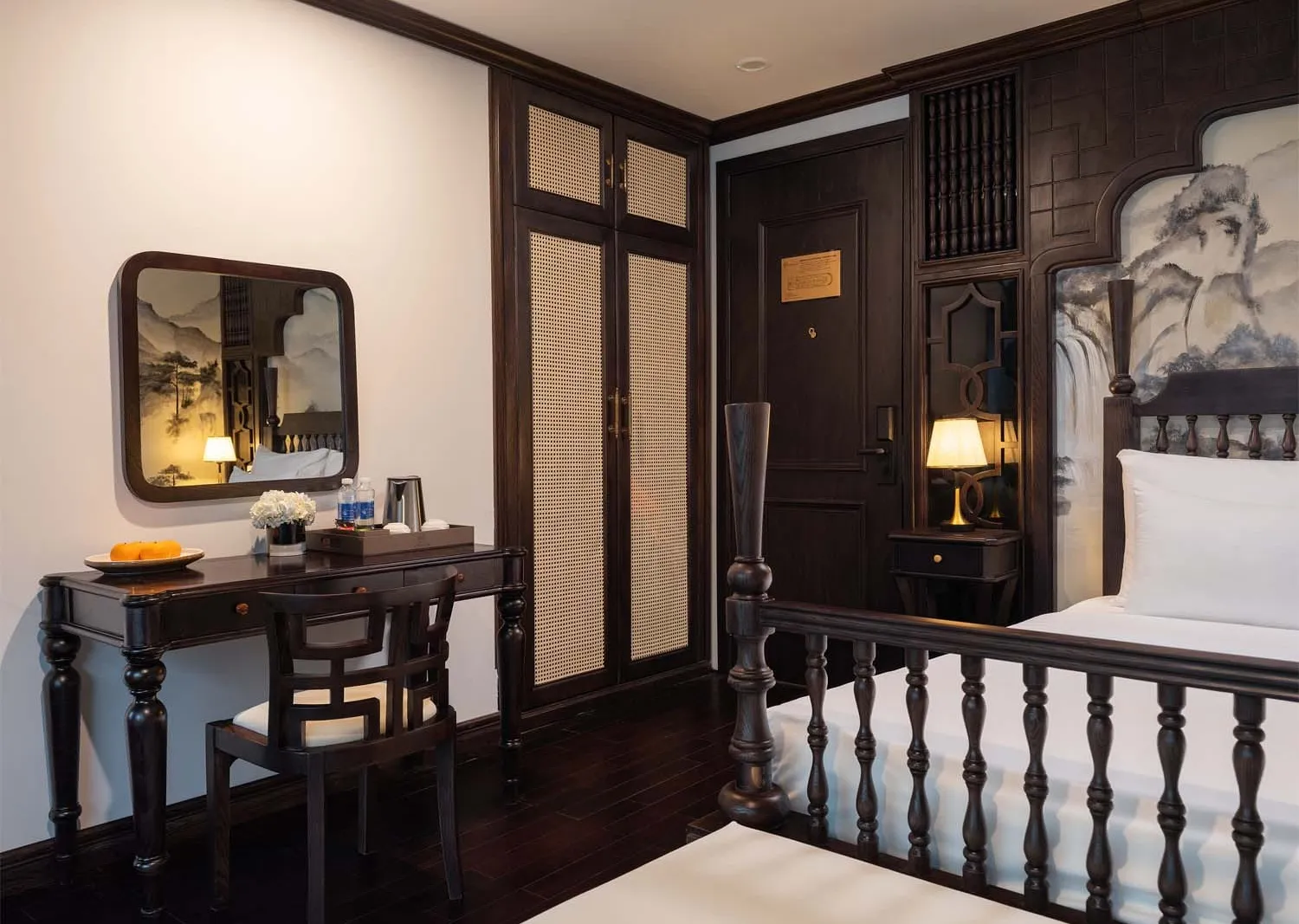 A dark-wood-furnished cruise cabin with a desk, mirror, wardrobe, bedside lamp, and a bed with white linens, decorated with ink-wash landscape artwork on the walls and headboard panel.