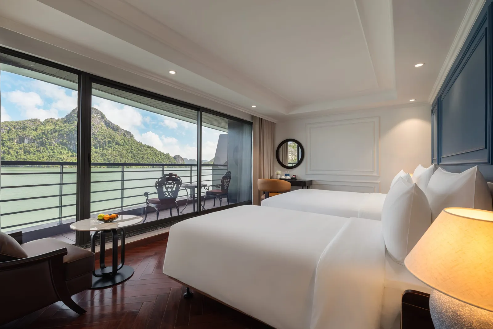 A luxury cabin interior with a white bed, dark wood flooring, and large windows showing limestone karsts and green water outside.