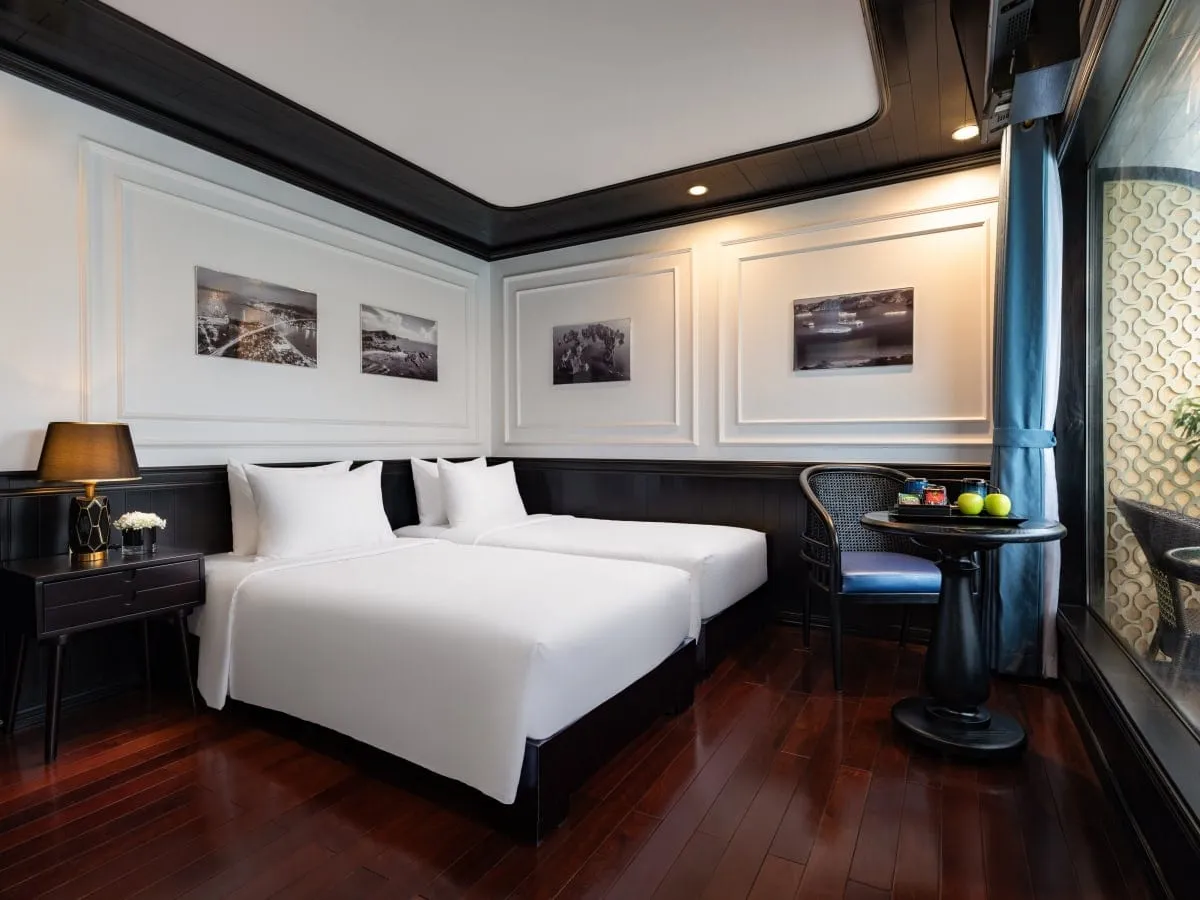 A cabin with two twin beds in white linen, dark wood floor and trim, a bedside lamp, a small round table with fruit and cups, and black-and-white bay photos on the wall.