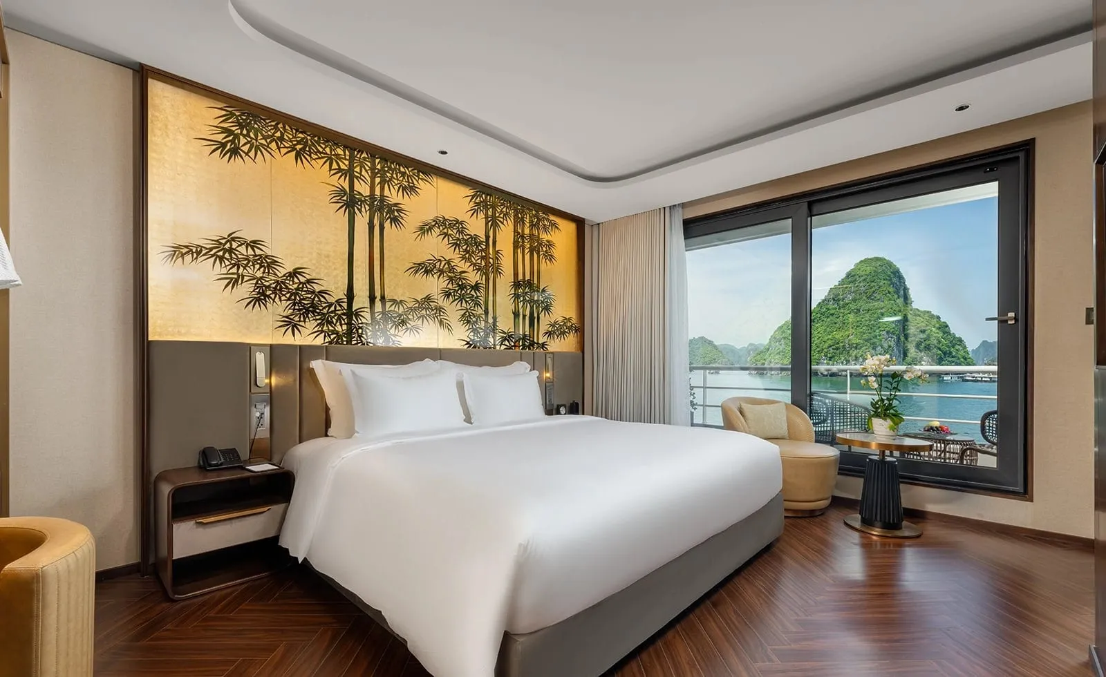 A cabin with a white king bed, a gold bamboo-painted headboard panel and herringbone wood floor, with floor-to-ceiling glass doors opening to a balcony and a view of green limestone karsts over the water.