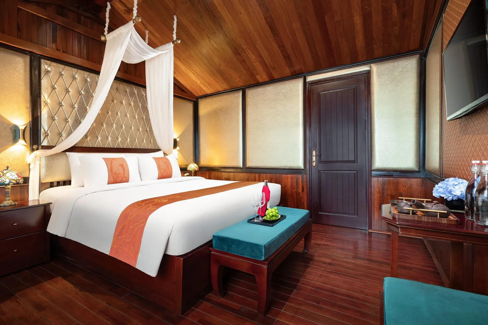 A wood-paneled cabin with a king bed under a white canopy, orange bed runner, teal bench with a fruit tray and wine glasses, a dark wooden door, a wall-mounted TV and a desk with water bottles and blue flowers.
