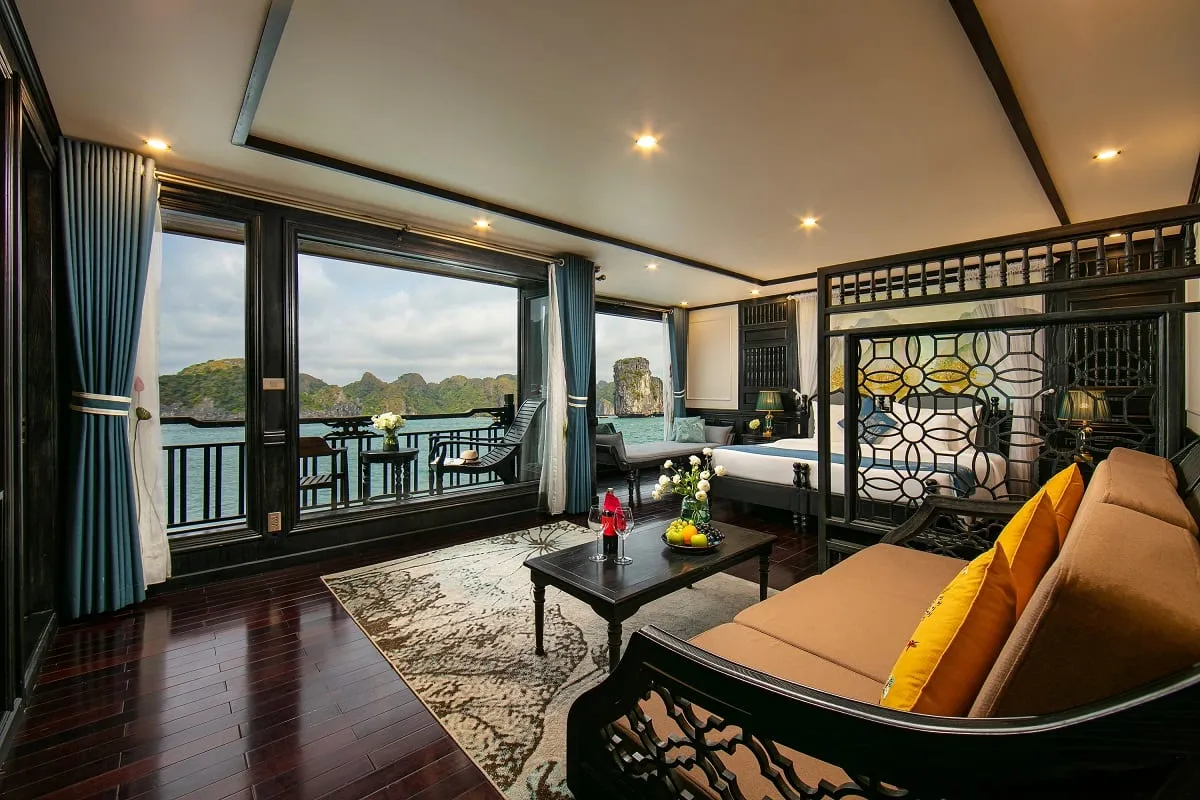 A luxury cruise cabin interior with dark wood floors, orange sofa, bed, and large windows overlooking limestone karsts and turquoise water.