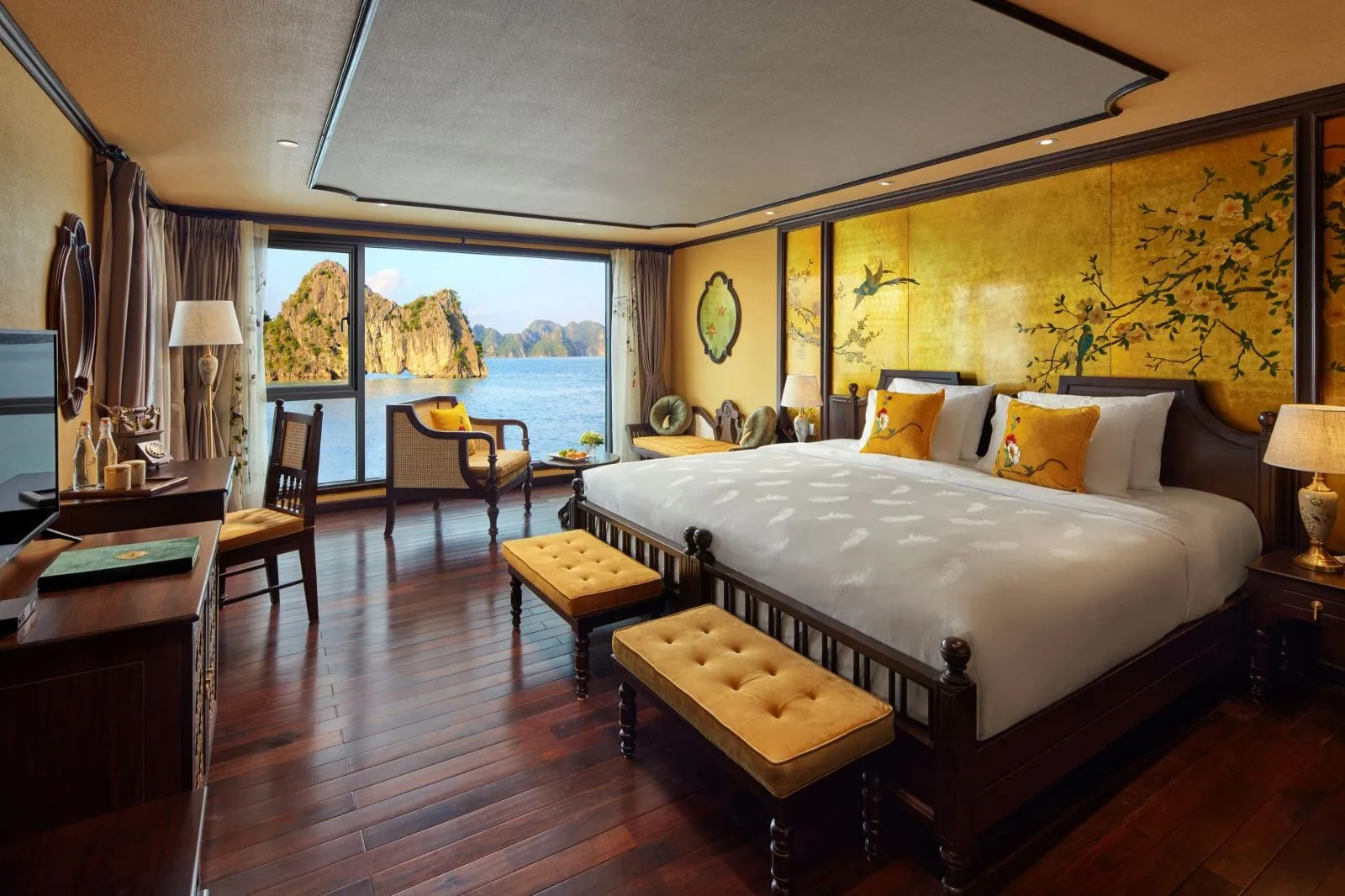A cruise cabin with a large bed, gold painted floral wall panels, yellow cushions and benches, dark wood floors, and a window looking out on limestone karsts rising from calm water.
