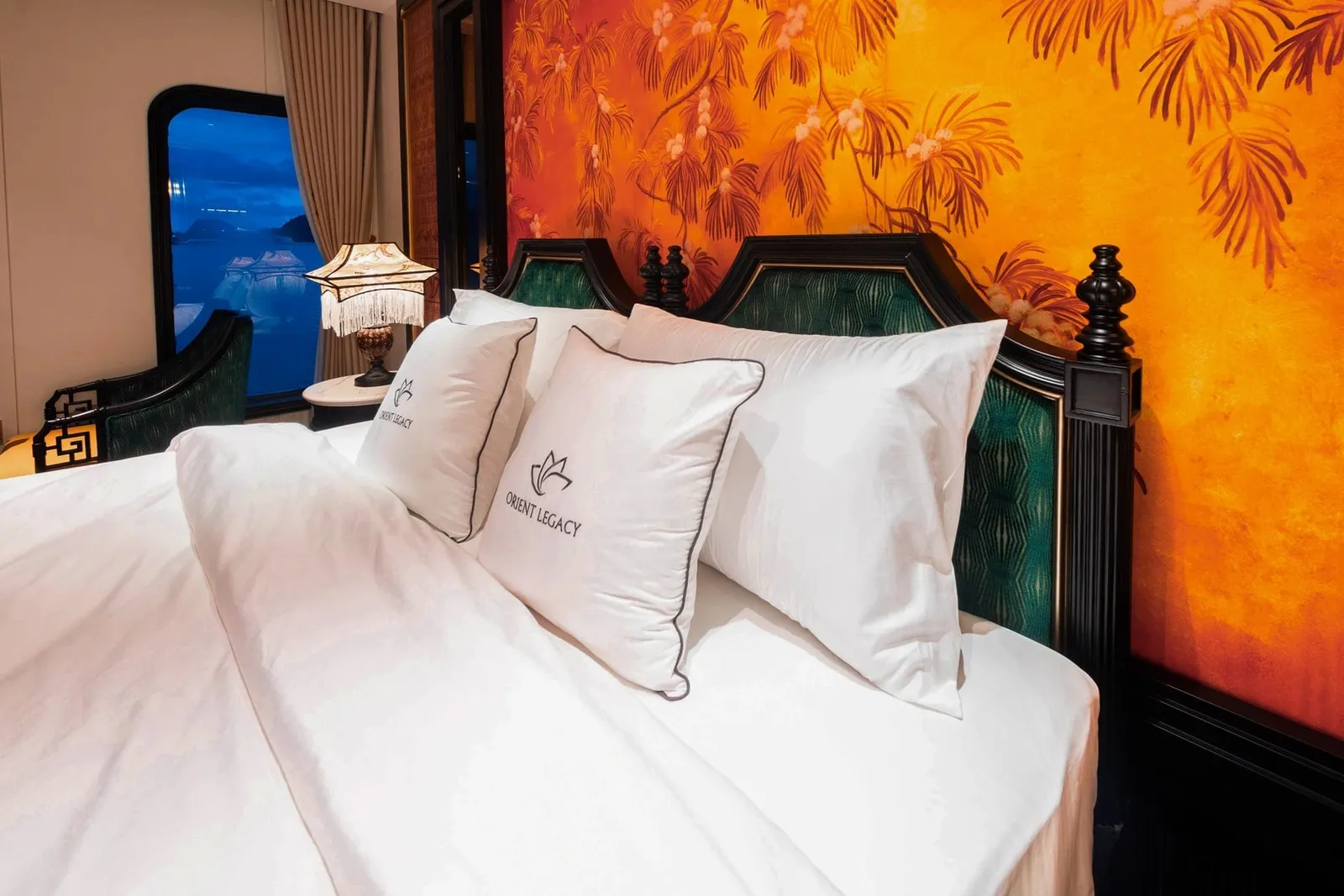 A cabin bed with white linens and cushions embroidered 'Orient Legacy', a dark wood green-velvet headboard, an orange floral wall mural, a fringed lamp, and a window showing blue water and distant karst islands.