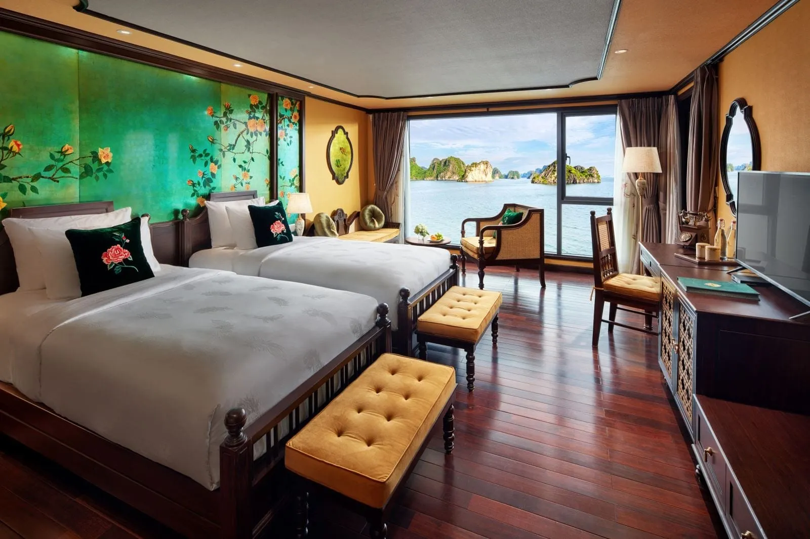 A cabin with two twin beds, gold tufted benches, a floral green wall panel and dark wood floor, with a large window showing limestone karst islands on calm water.