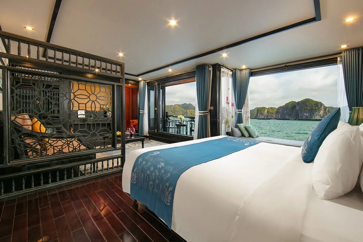 A luxury cruise cabin interior with white bedding, decorative screens, and large windows overlooking limestone karsts and turquoise water.