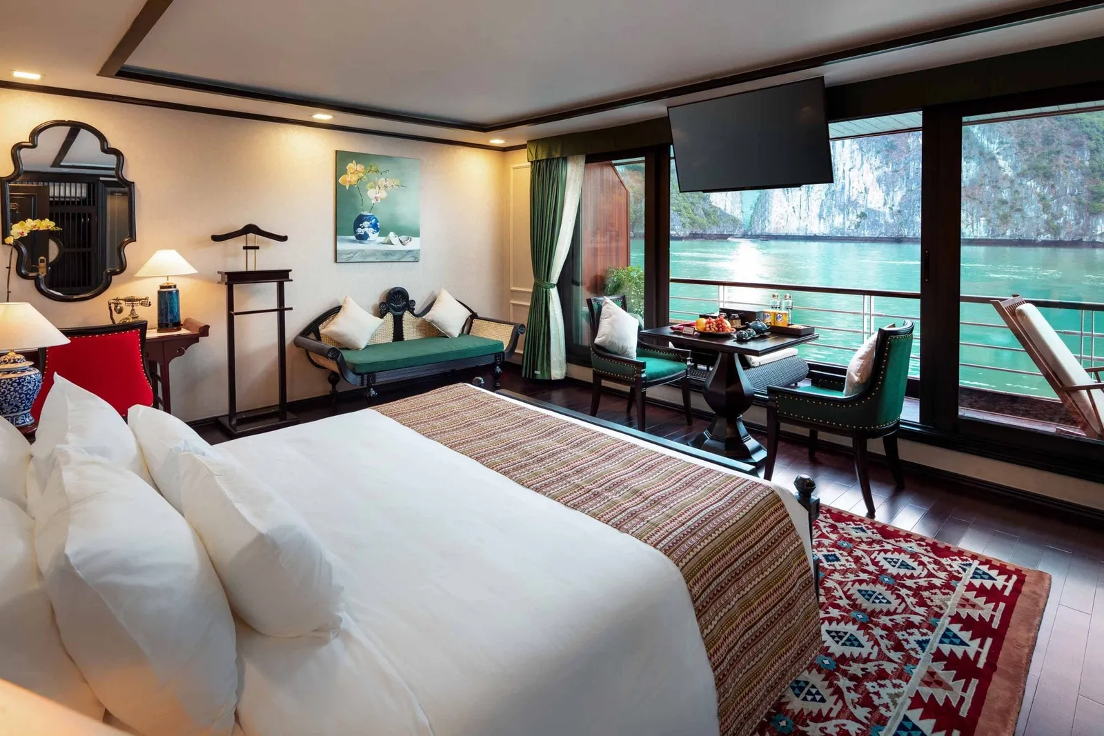 A cabin with a large white bed, green velvet chairs, a bench and a wall-mounted TV, with floor-to-ceiling windows and a balcony overlooking turquoise water and limestone karsts.