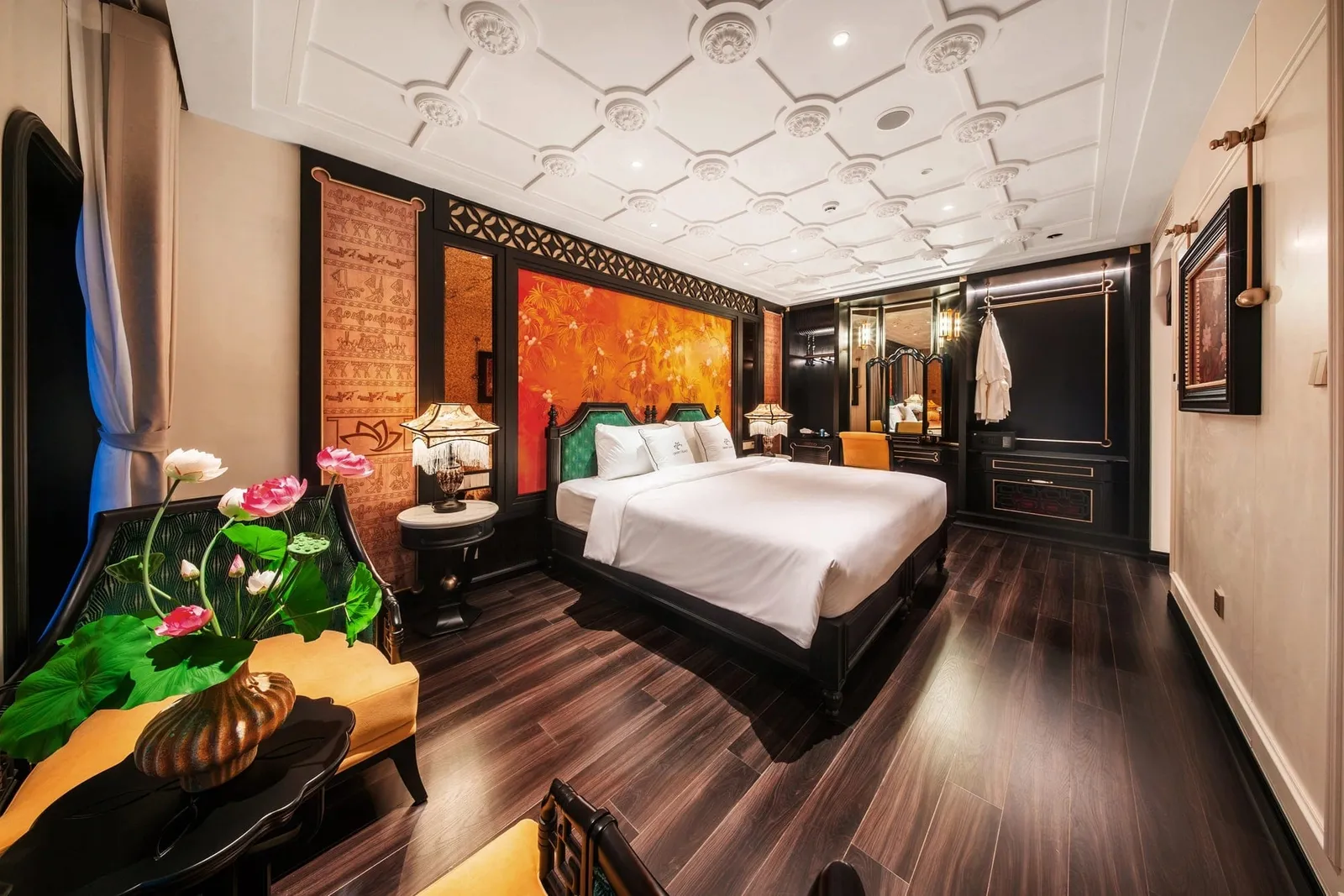 A luxurious cabin with a large white-draped bed, dark wood floor, orange lacquer-style artwork behind the headboard, lotus flowers in a vase in the foreground, and a robe hanging in an open wardrobe area.