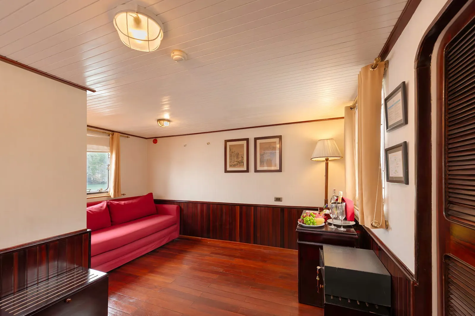 Interior of a wood-paneled cabin with a red sofa, a side table holding a fruit plate and champagne glasses, framed pictures on the walls, a table lamp, and a porthole window showing a rocky green shoreline and water.