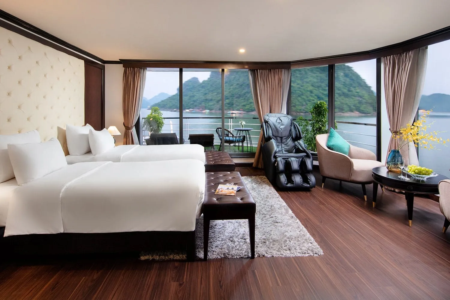 A cruise cabin with two twin beds, a massage chair, an armchair and a side table with fruit, with floor-to-ceiling windows and a balcony overlooking green limestone karsts on calm water.