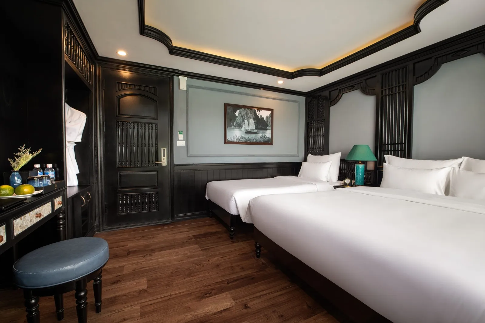 A twin-bed cabin interior with dark wood paneling, white bedding, wooden floors, and decorative Vietnamese-style carved trim.