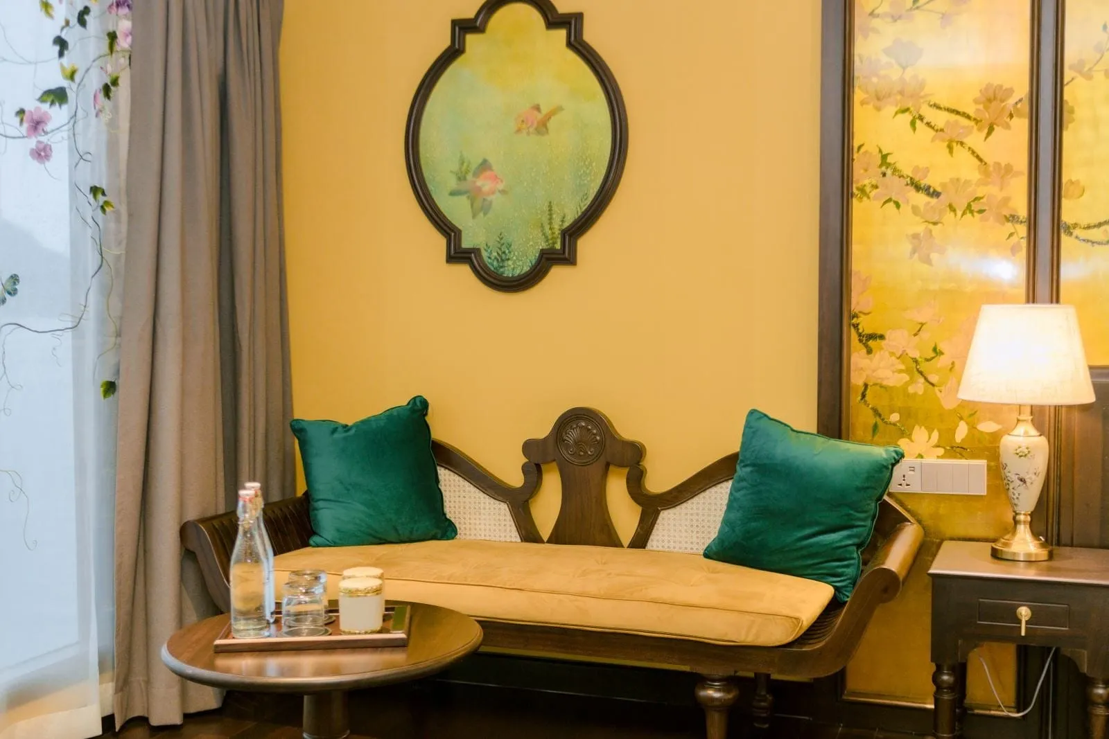A cabin sitting area with a carved wooden settee with a mustard cushion and two green velvet pillows, a round table holding water bottles and glasses, a framed goldfish painting on a yellow wall, a bedside table with a lamp, and floral curtains.