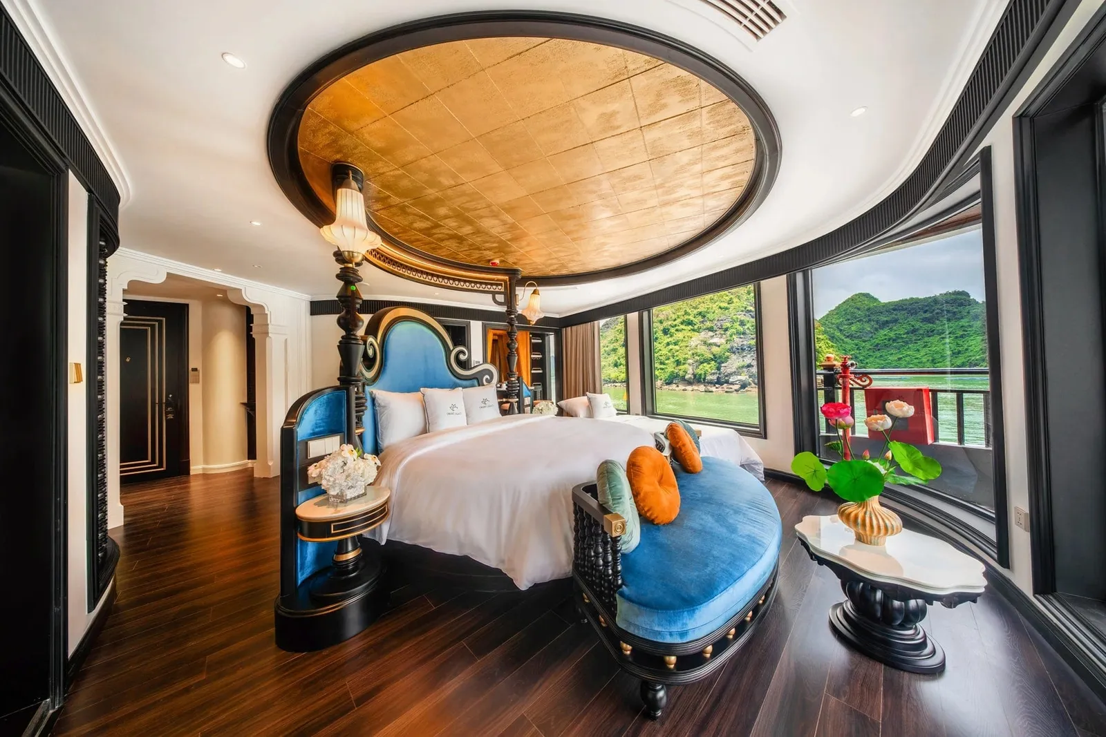 A luxurious cruise cabin with a gold-leaf oval ceiling, a white-linen bed with blue velvet headboard and bench, dark wood floors, and curved windows showing green limestone karst hills over the water; a small logo appears on the pillows.