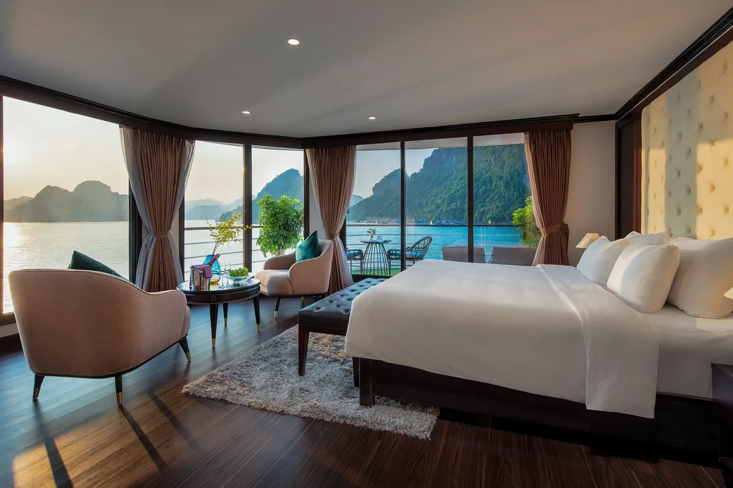A cabin interior with a large bed, armchairs and a round table, with floor-to-ceiling windows and a balcony overlooking calm water and limestone karst islands at sunset.