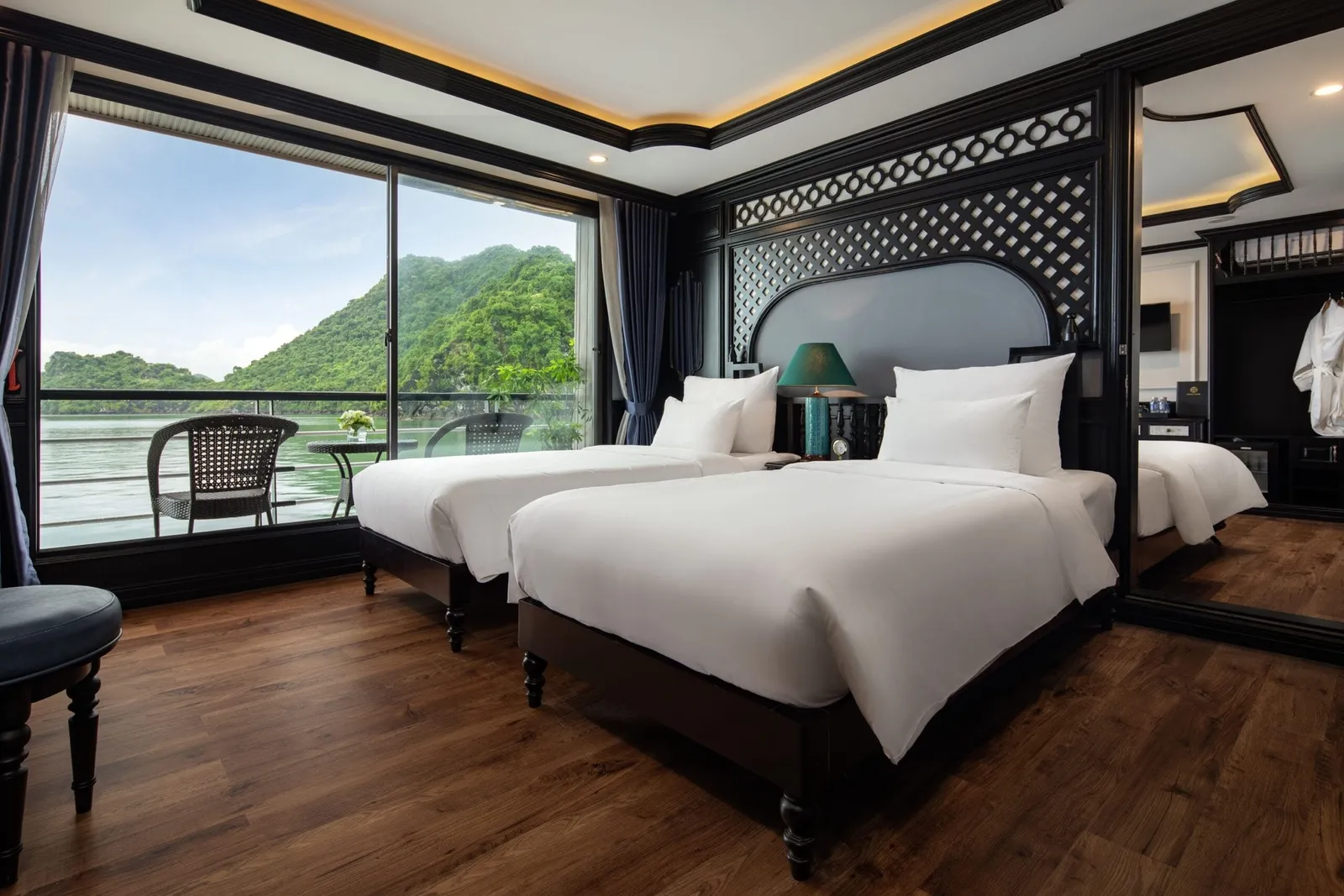 A cruise boat cabin with two beds, decorative black headboard, and large windows showing green karst mountains and calm water outside.