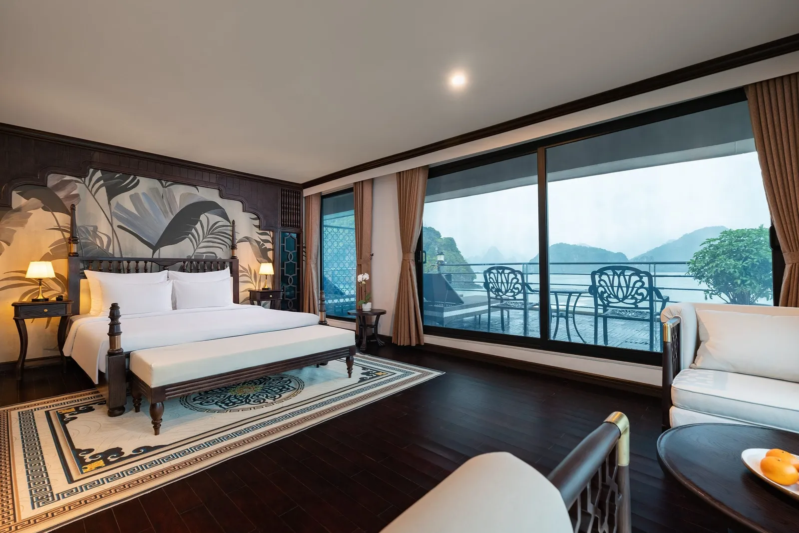 A spacious cabin with a white-bedded double bed, dark wood floor, sofa and a private balcony with chairs, looking out onto misty limestone karsts.