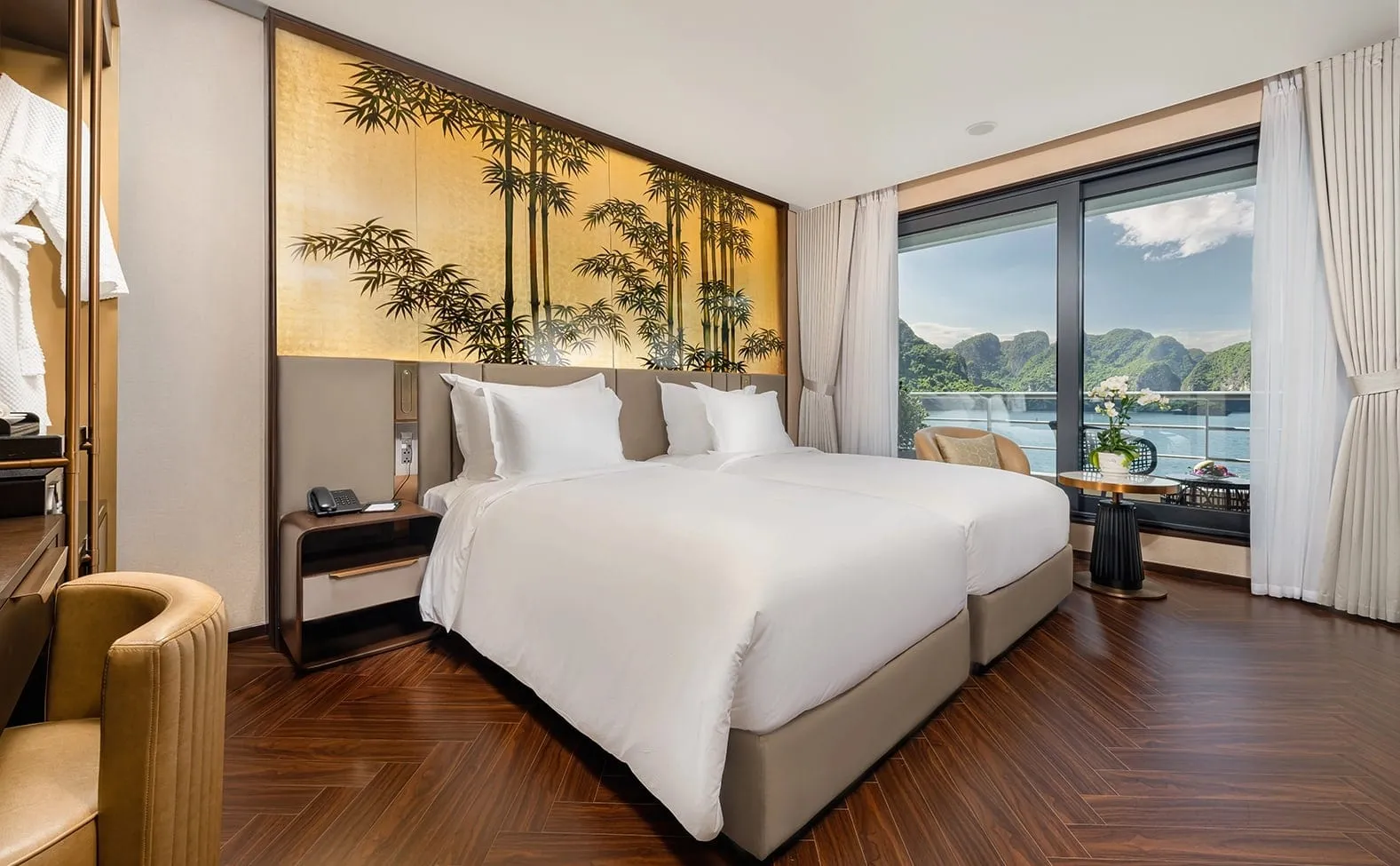 A cabin with two white-linen twin beds, a bamboo-painted gold headboard panel, herringbone wood floor, and a window and balcony looking out on limestone karst islands and the sea.