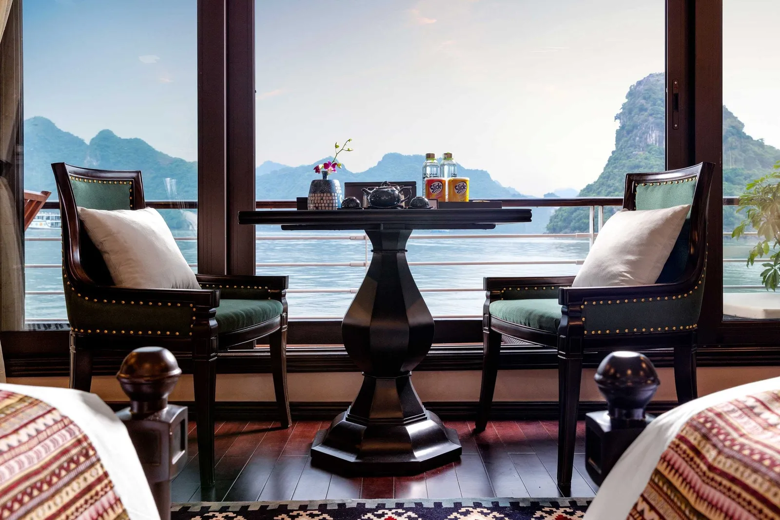 Cabin interior with a dark wooden table and two green upholstered chairs beside floor-to-ceiling windows overlooking limestone karst peaks and calm water, with bed ends in the foreground.